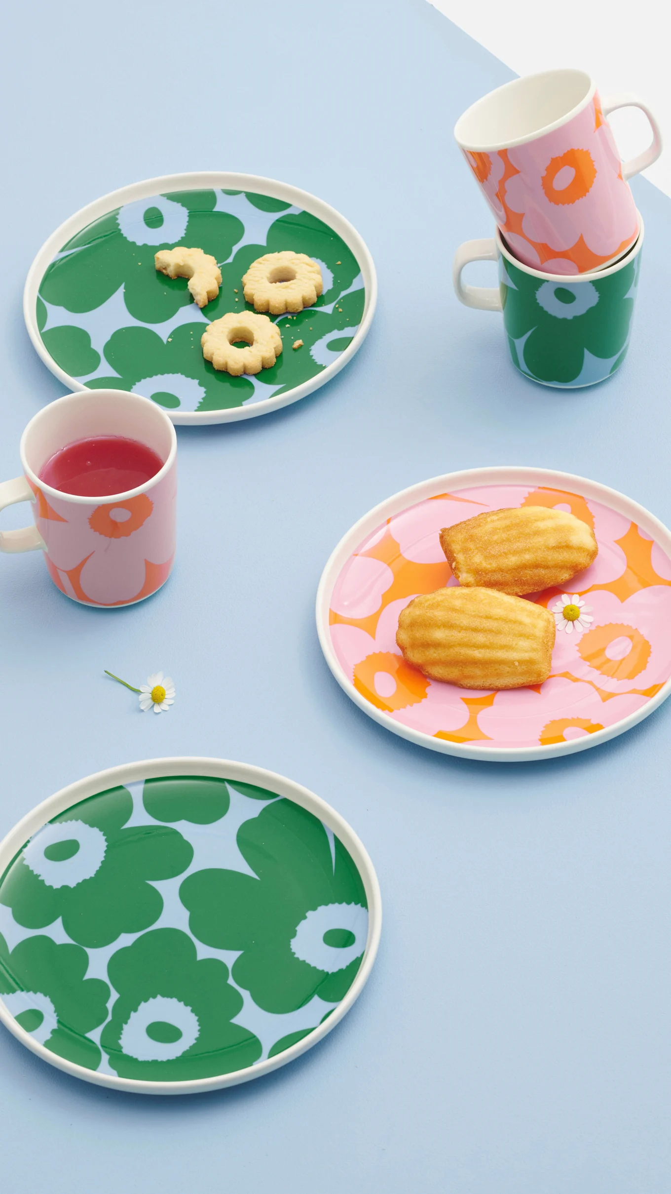 Assorted floral patterned plates and mugs with cookies, madeleines, and a red drink on a light blue background.