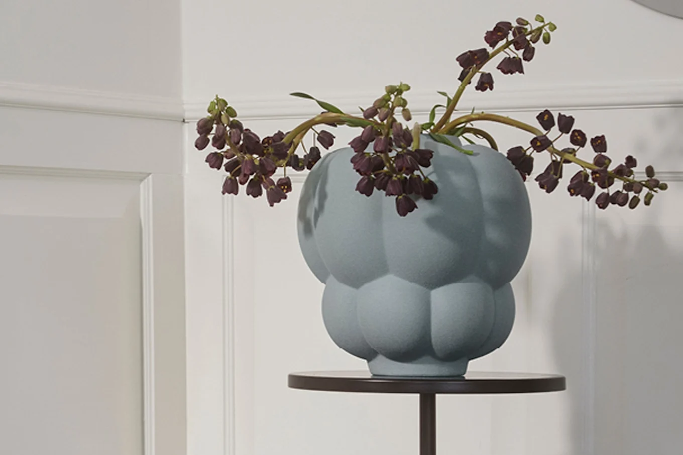 A light blue bulbous vase with dark purple bell-shaped flowers on a dark round table.