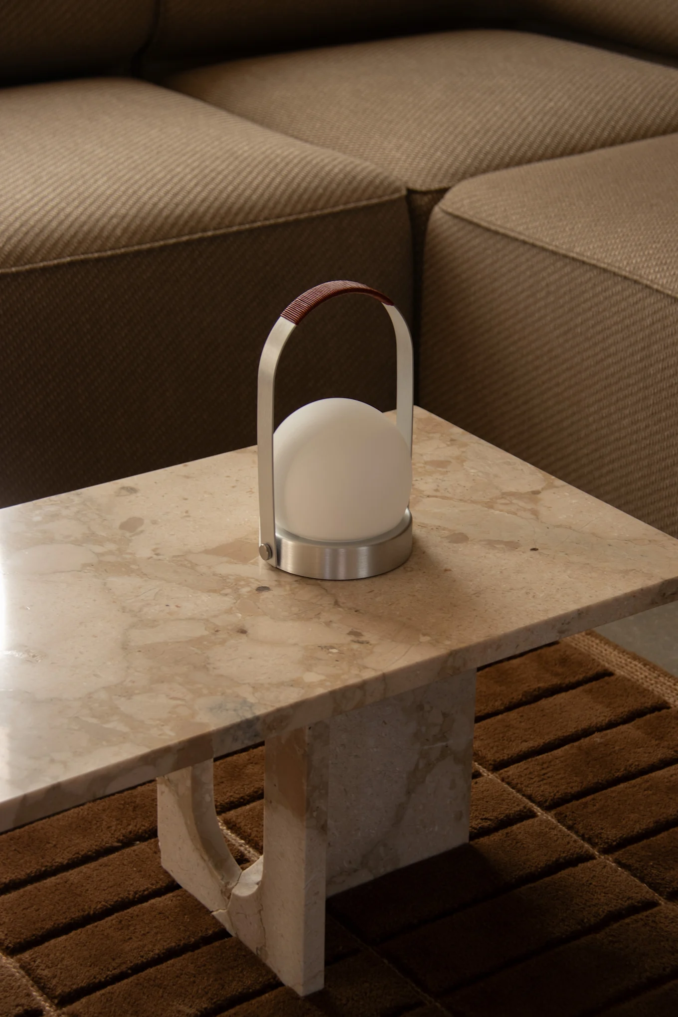 A modern portable lamp with a white spherical light, brushed metal frame, and brown leather handle sits on a light marble table.