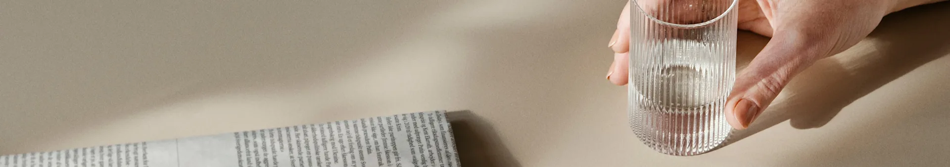 A hand holds a ribbed glass of water next to a rolled newspaper on a light background.