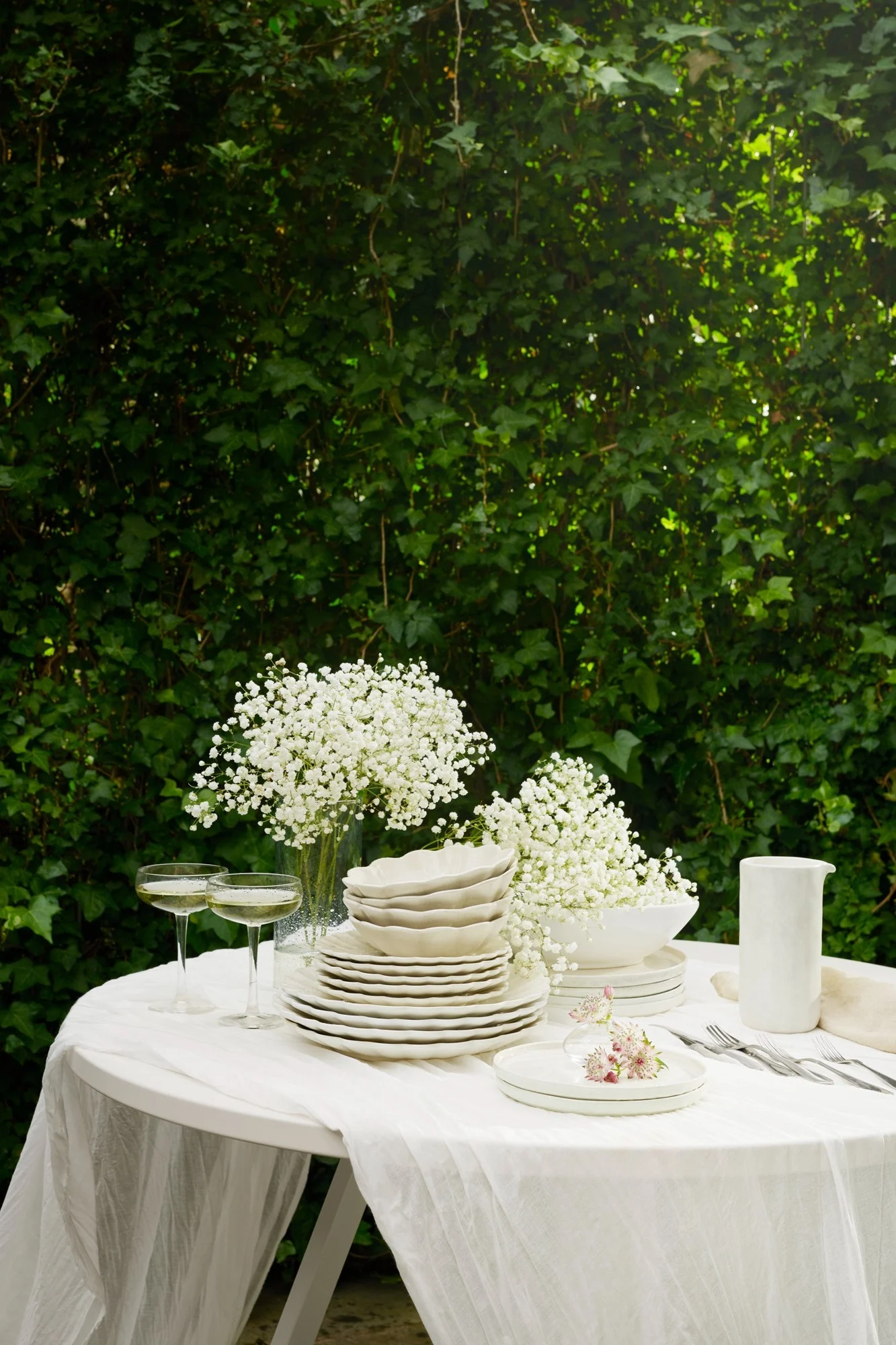 Outdoor table set with white dinnerware, baby's breath flowers, champagne flutes, and silverware on a white tablecloth, against lush green leaves.