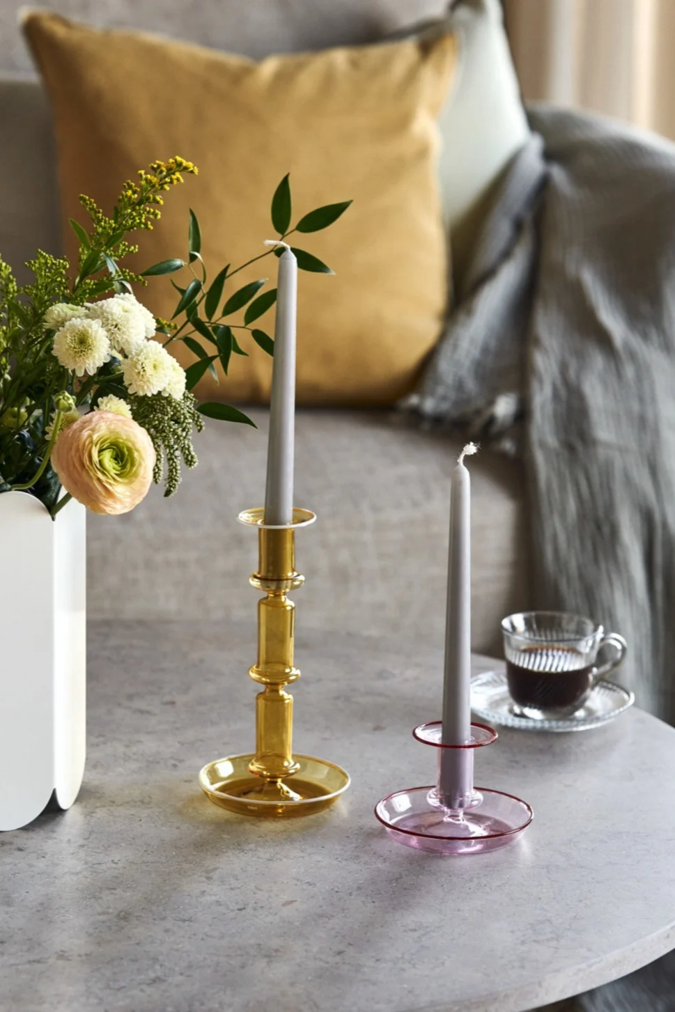Two glass candlesticks, one amber and one pink, with grey candles on a table next to a vase of flowers and a cup of coffee.
