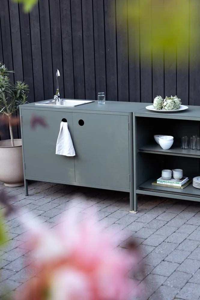 Outdoor kitchen cabinet in dark green with a sink, faucet, and open shelves filled with dishes and artichokes.