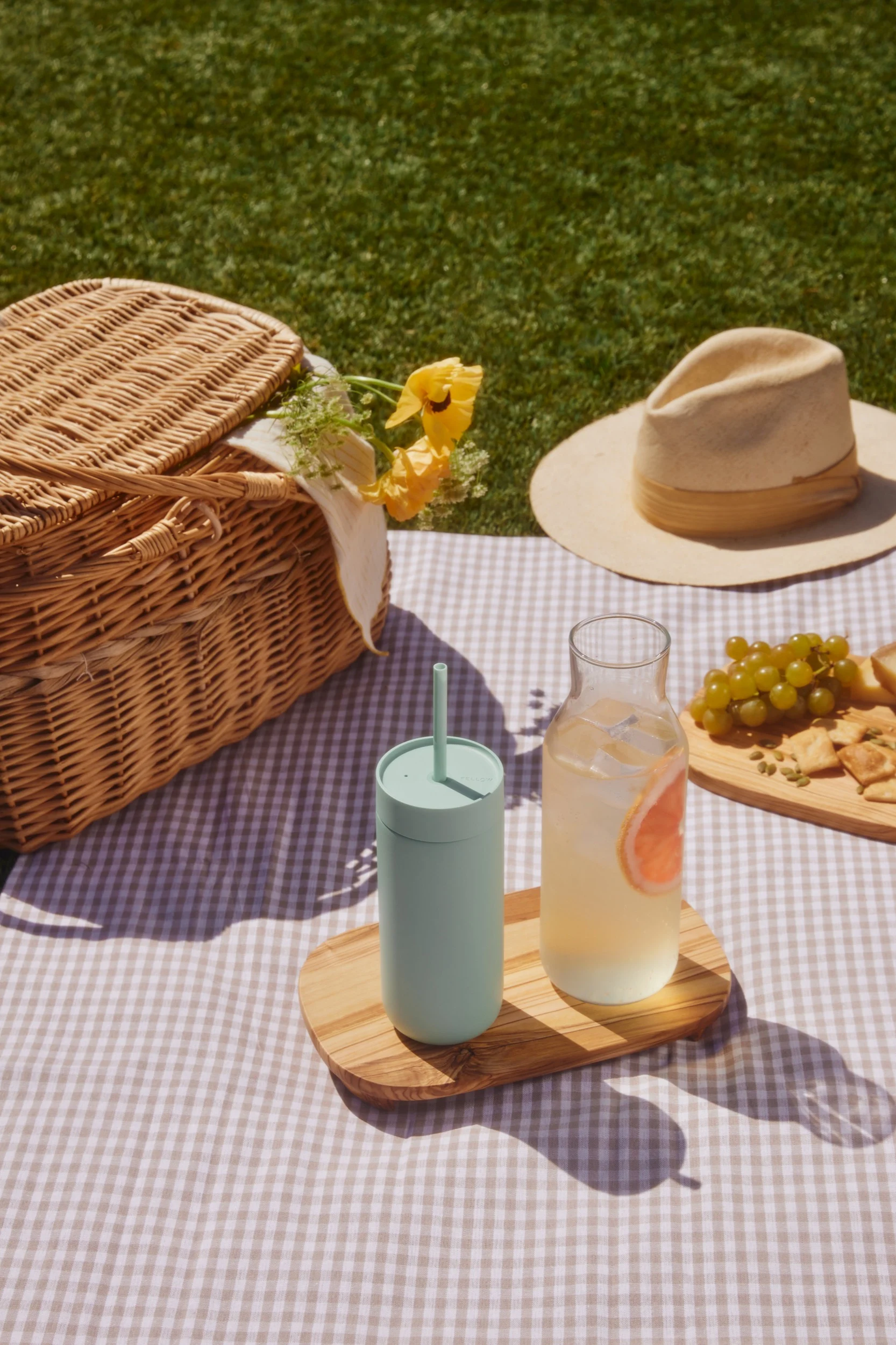 Picnic on a checkered blanket: wicker basket, light green tumbler, grapefruit drink, grapes, and crackers on grass.