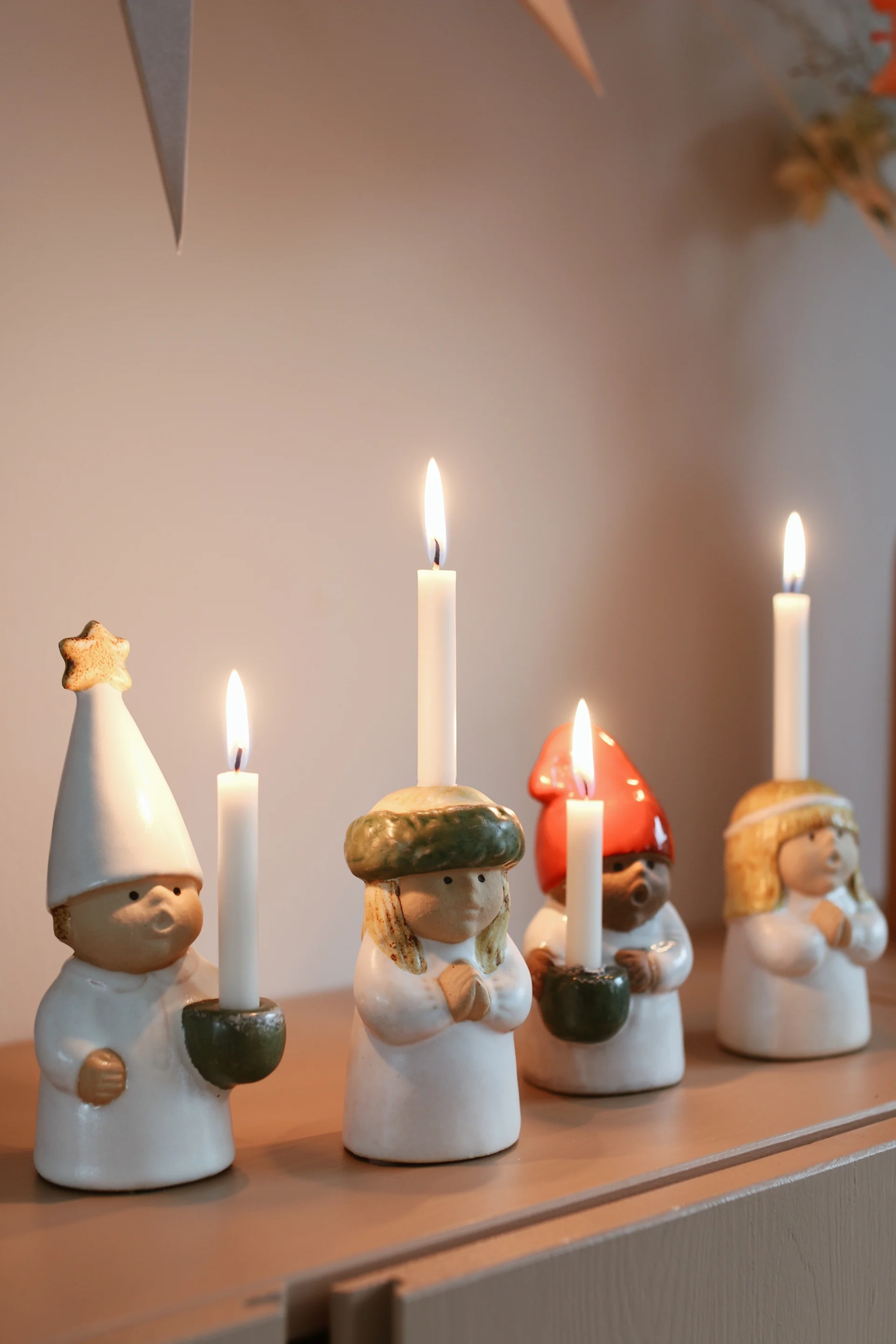 Four ceramic figures with diverse hats and hairstyles, each holding a lit white candle on a wooden surface.
