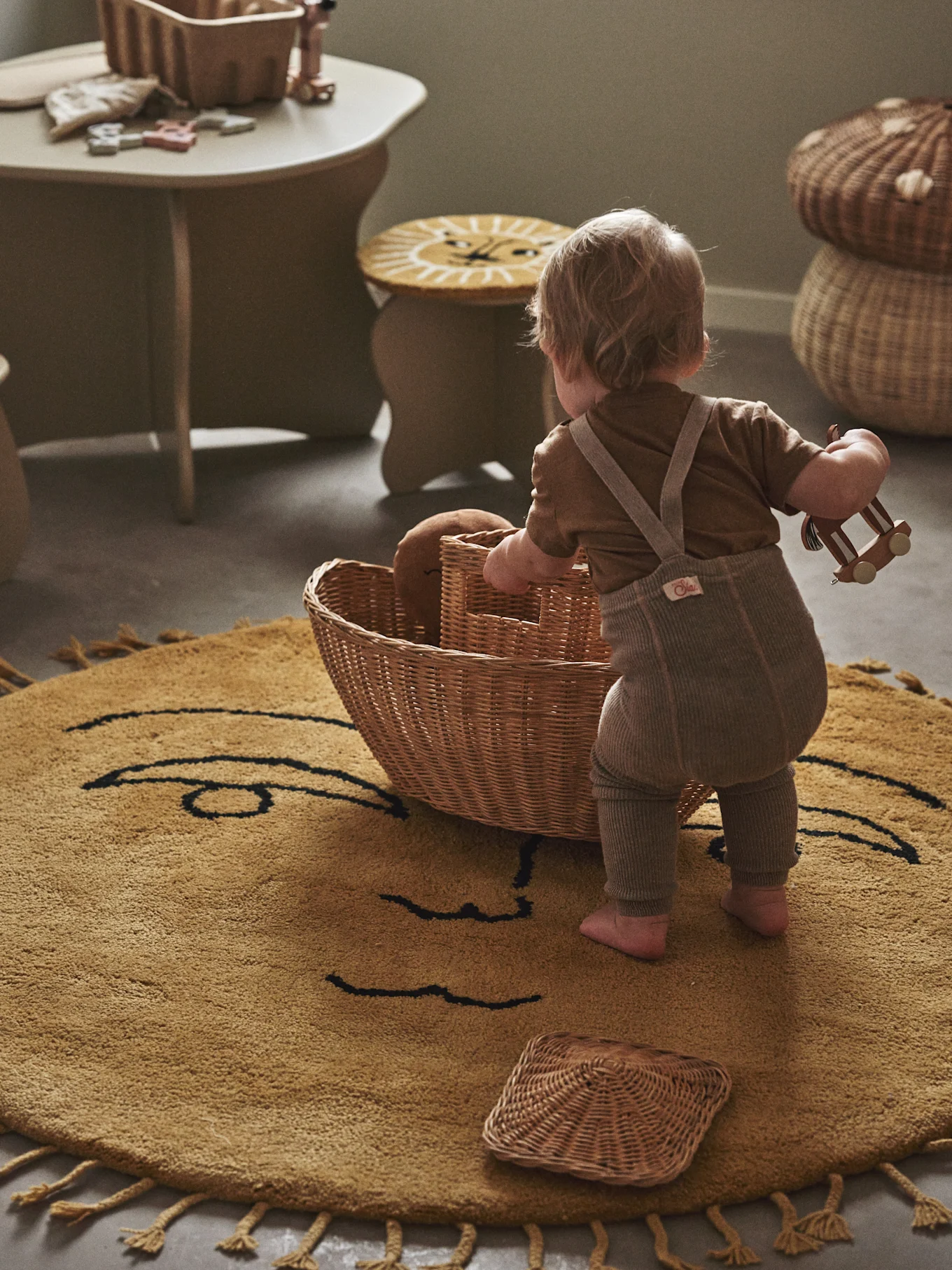 Baby with a wooden toy car and wicker basket on a round, yellow rug with a smiling face design.