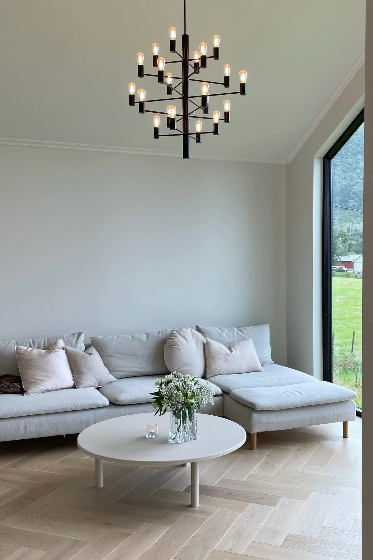 A modern living room with a black chandelier, grey sectional sofa, round coffee table with flowers, and a large window overlooking a green landscape.