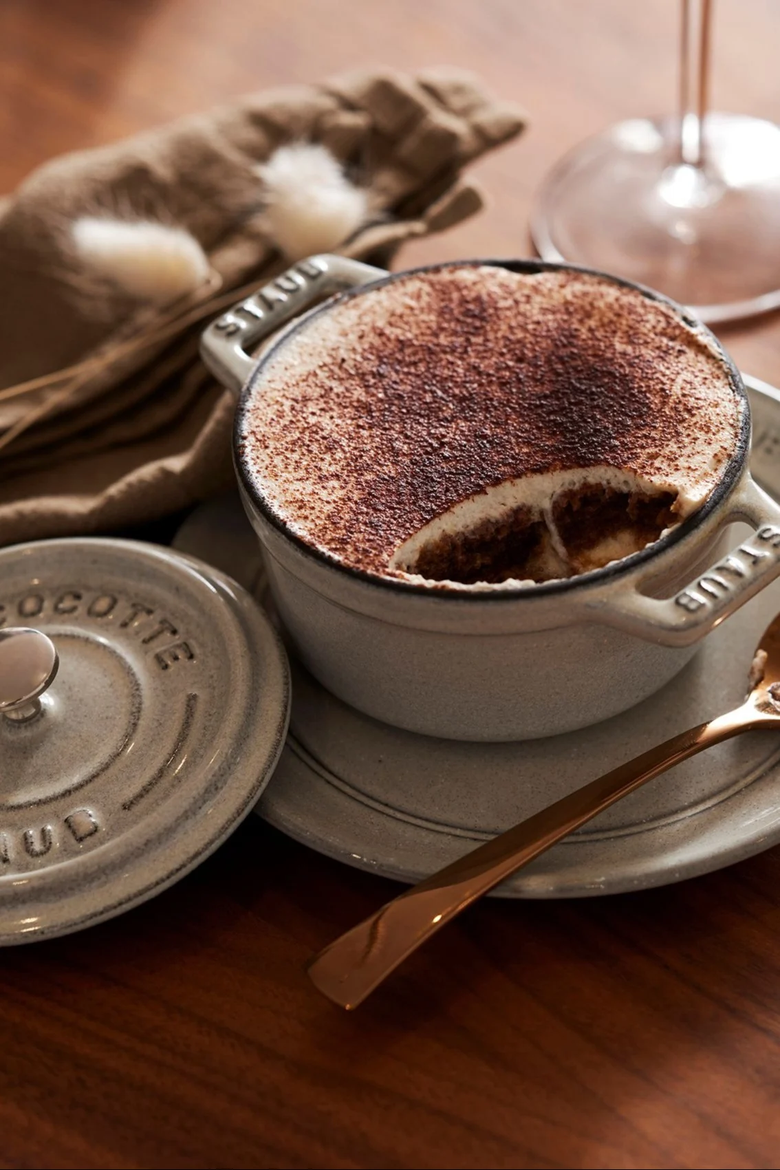 A grey cast iron mini cocotte filled with tiramisu, topped with cocoa powder, with a scoop taken out. A golden spoon and the cocotte lid are beside it on a wooden table.