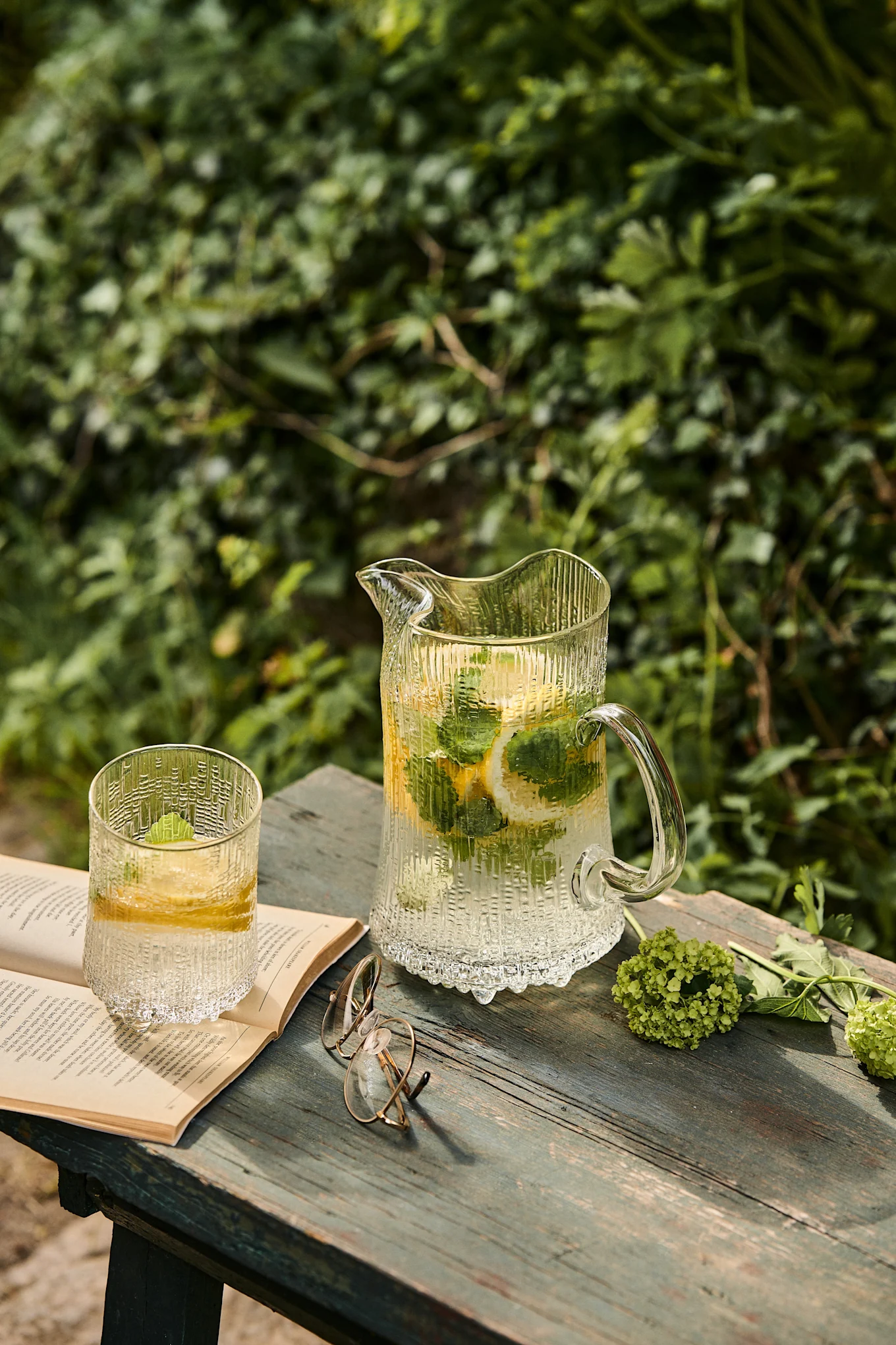 Refreshing lemonade with lemon and mint in a textured glass pitcher and drinking glass on a wooden table with an open book and spectacles.