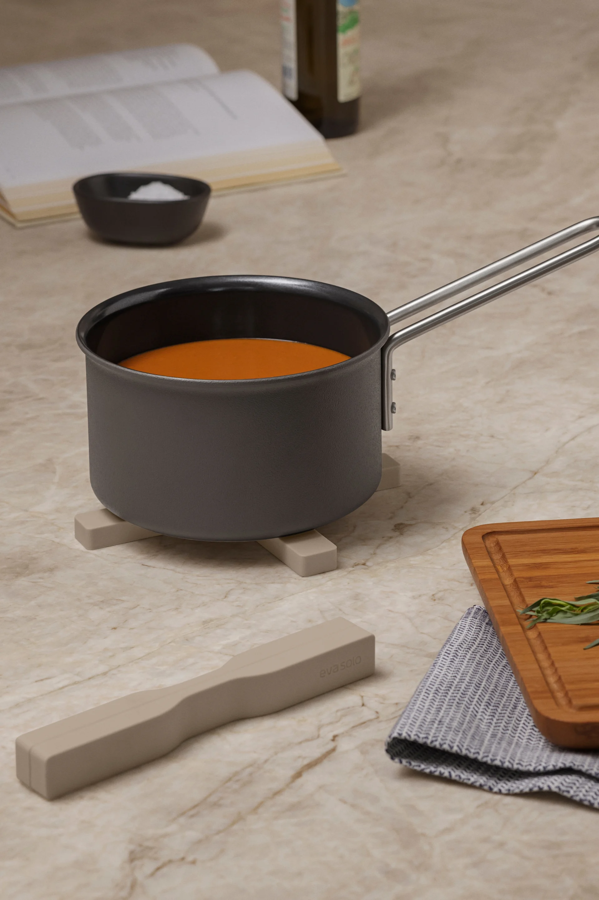Saucepan with orange liquid on a trivet, with a cookbook, salt, whisk, herbs on a cutting board, and a utensil on a marble counter.