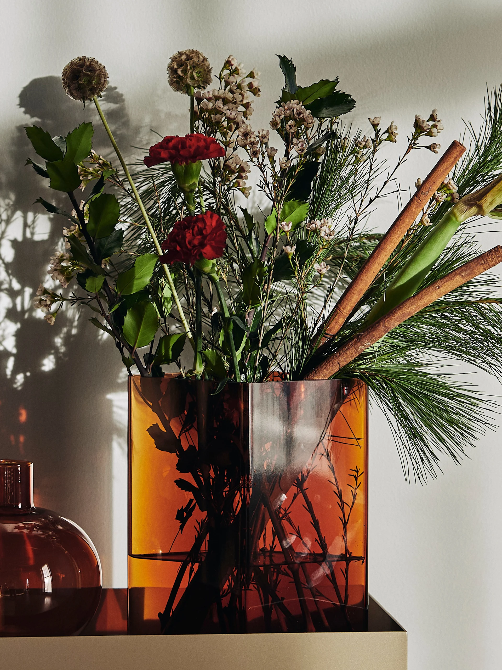 Festive bouquet in an amber square vase with red carnations, pine needles, cinnamon sticks, and small white flowers.