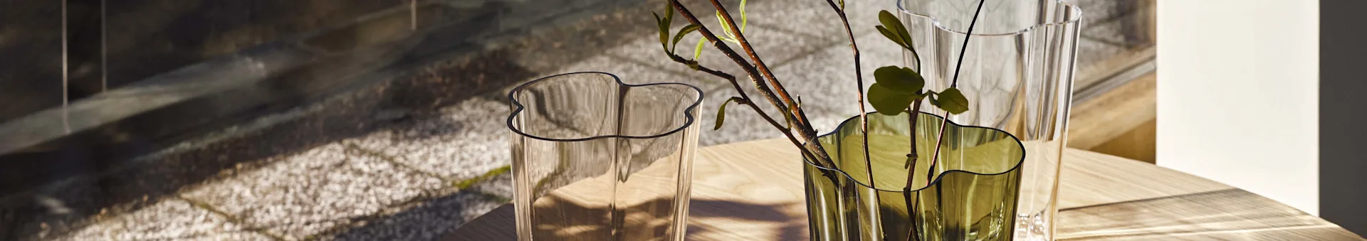 Two wavy glass vases, one clear and one green with branches, sit on a light wooden table with shadows.