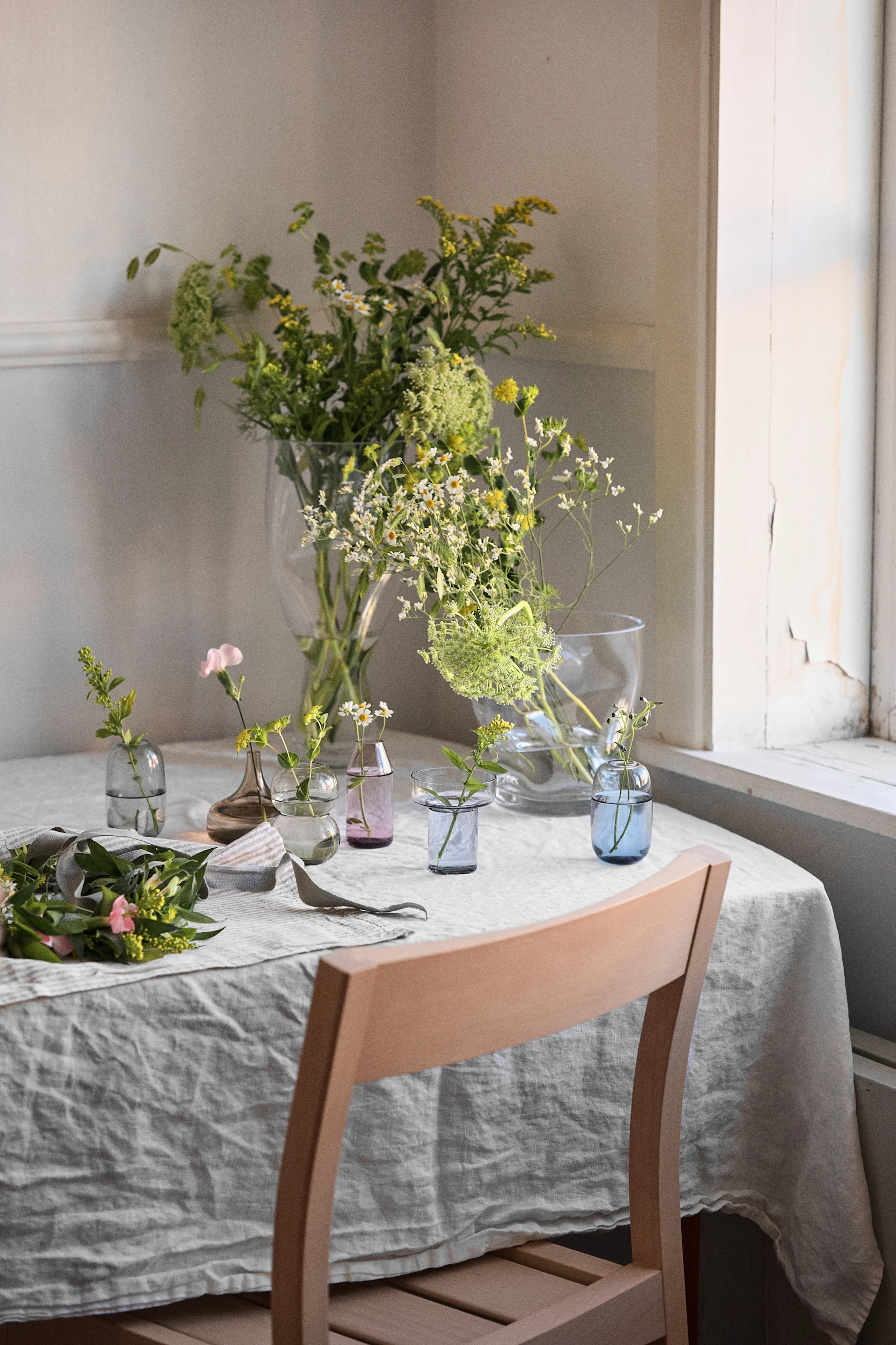 Table with linen, various vases with green, yellow, and pink flowers. Wooden chair partially visible.