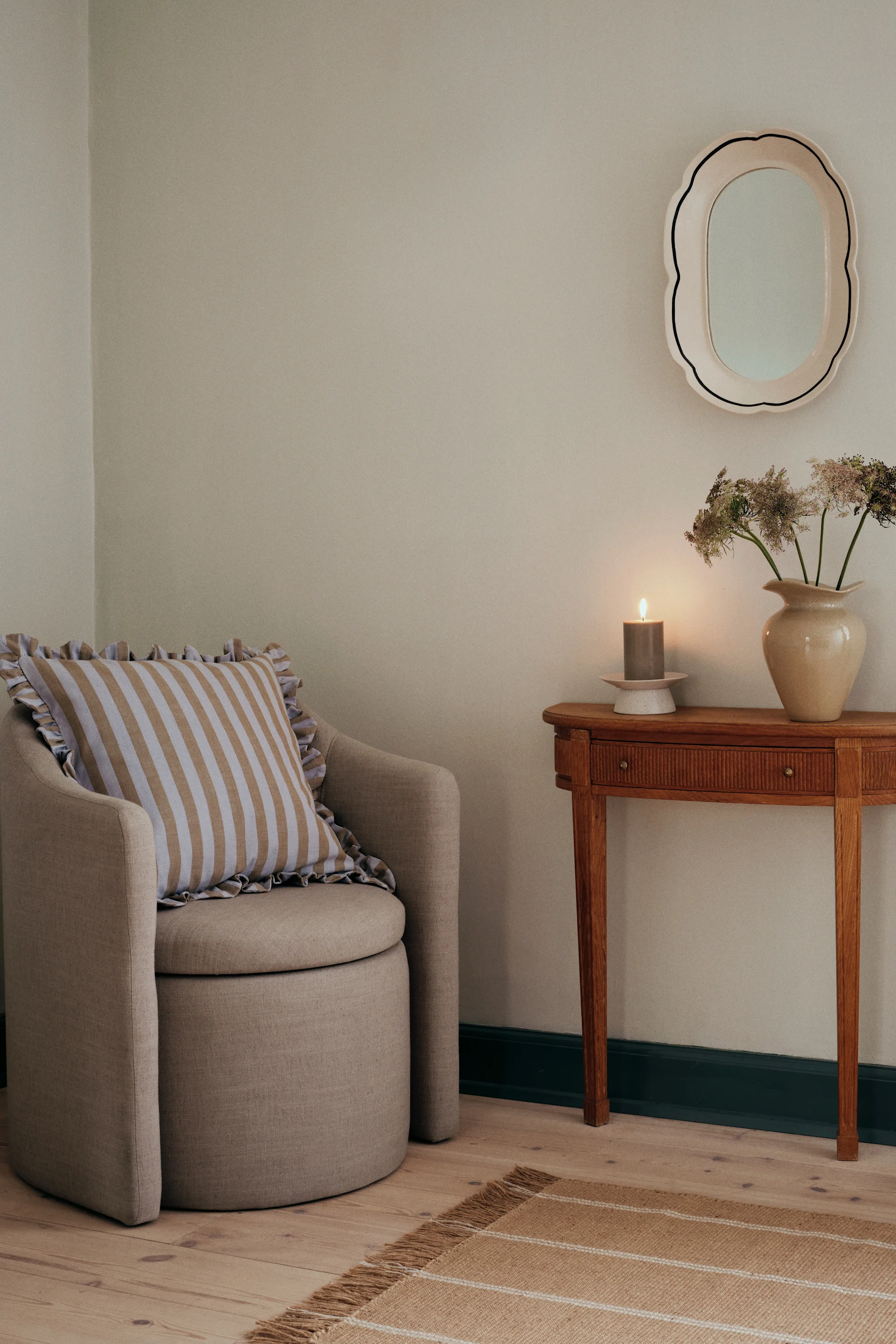 An armchair with a striped cushion, a wooden side table with a lit candle and vase of flowers, a mirror, and a striped rug.
