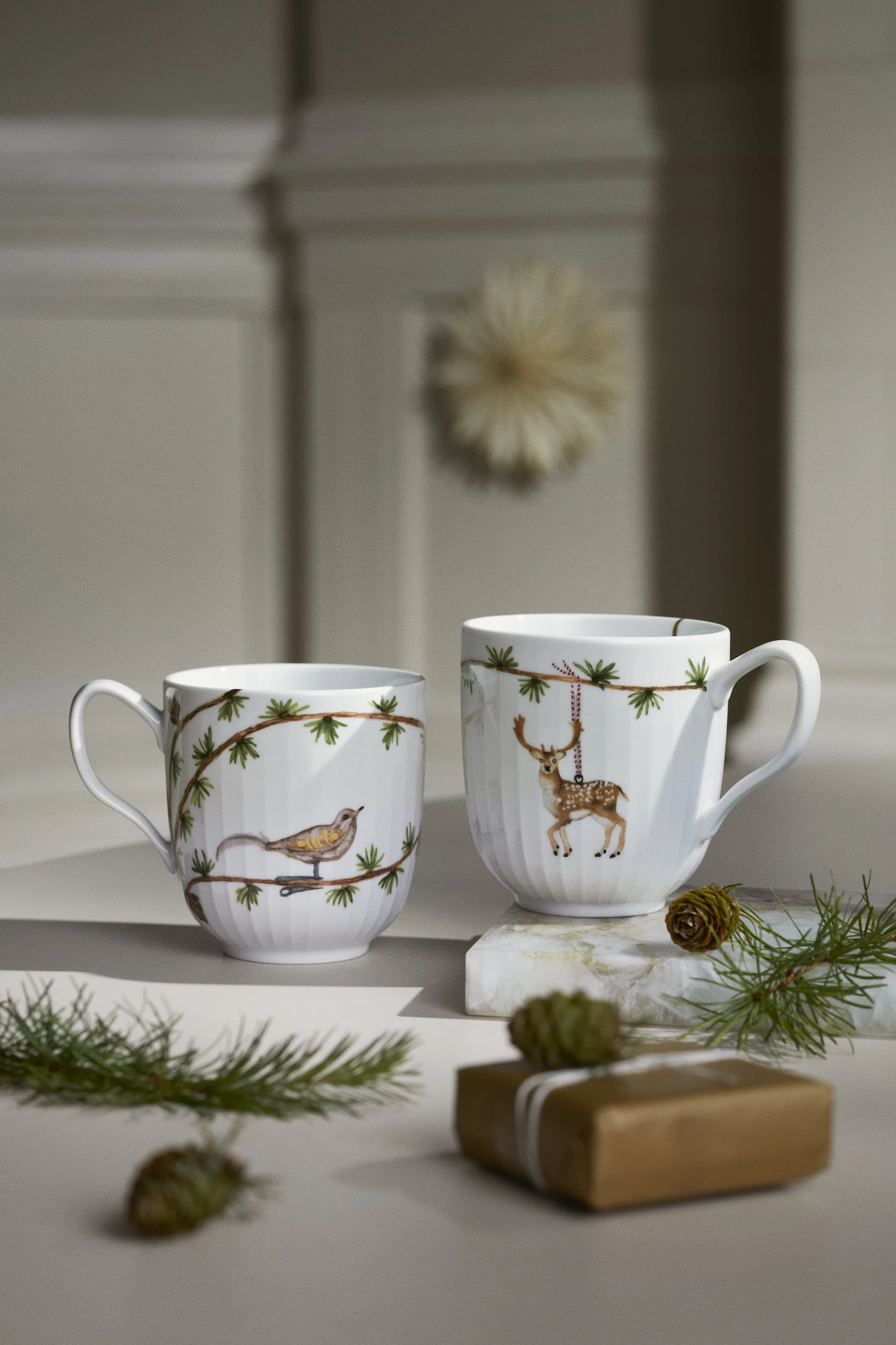 Two white mugs with nature-inspired holiday designs: a bird on a pine branch and a hanging spotted deer, with pine needles and a gift.