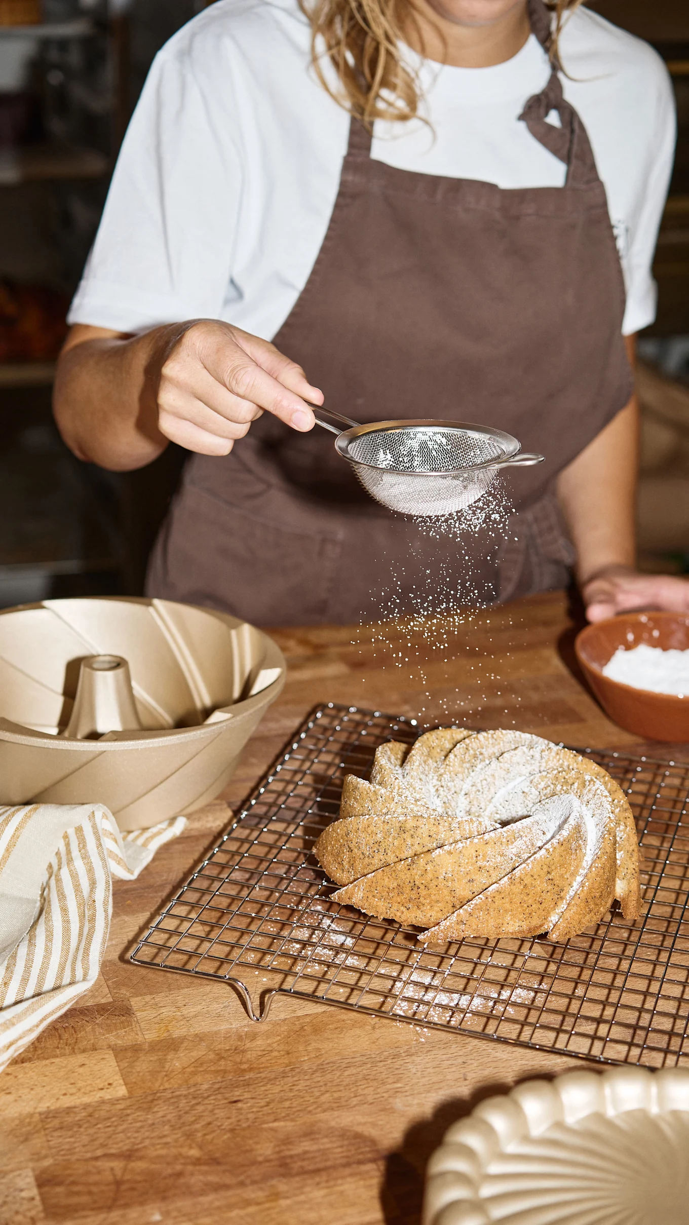 Hands sift powdered sugar over a bundt cake on a cooling rack, with a bundt pan nearby.