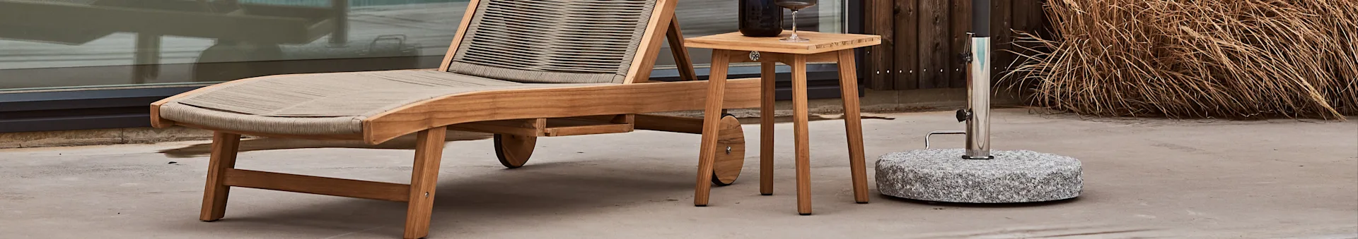 Outdoor scene with a wooden chaise lounge, a small side table with a drink, and a granite umbrella base.