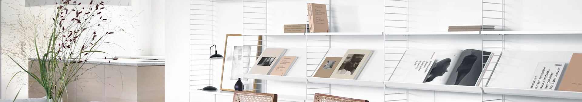 A white modular shelving system against a white wall, filled with books and magazines. A tall plant stands to the left.