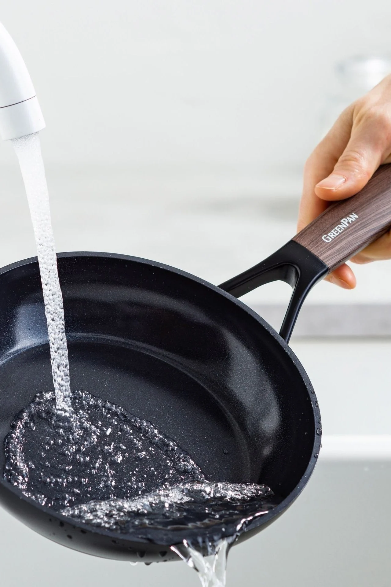 A hand holds a black frying pan with a wood-grain handle under a stream of running water in a sink.