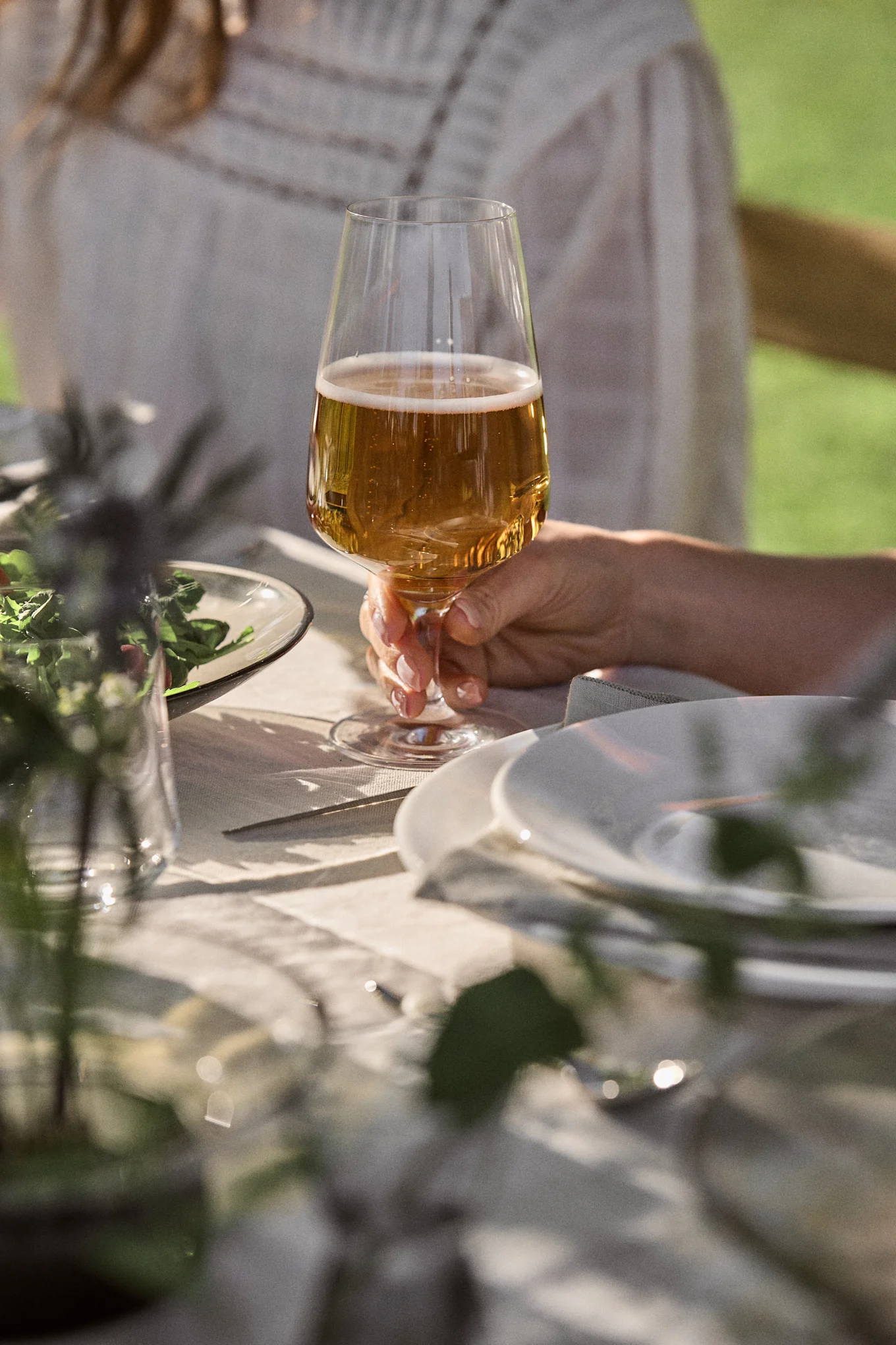 Hand holding a glass of beer with a foamy head at an outdoor table. Plates and greenery are blurred in the background.