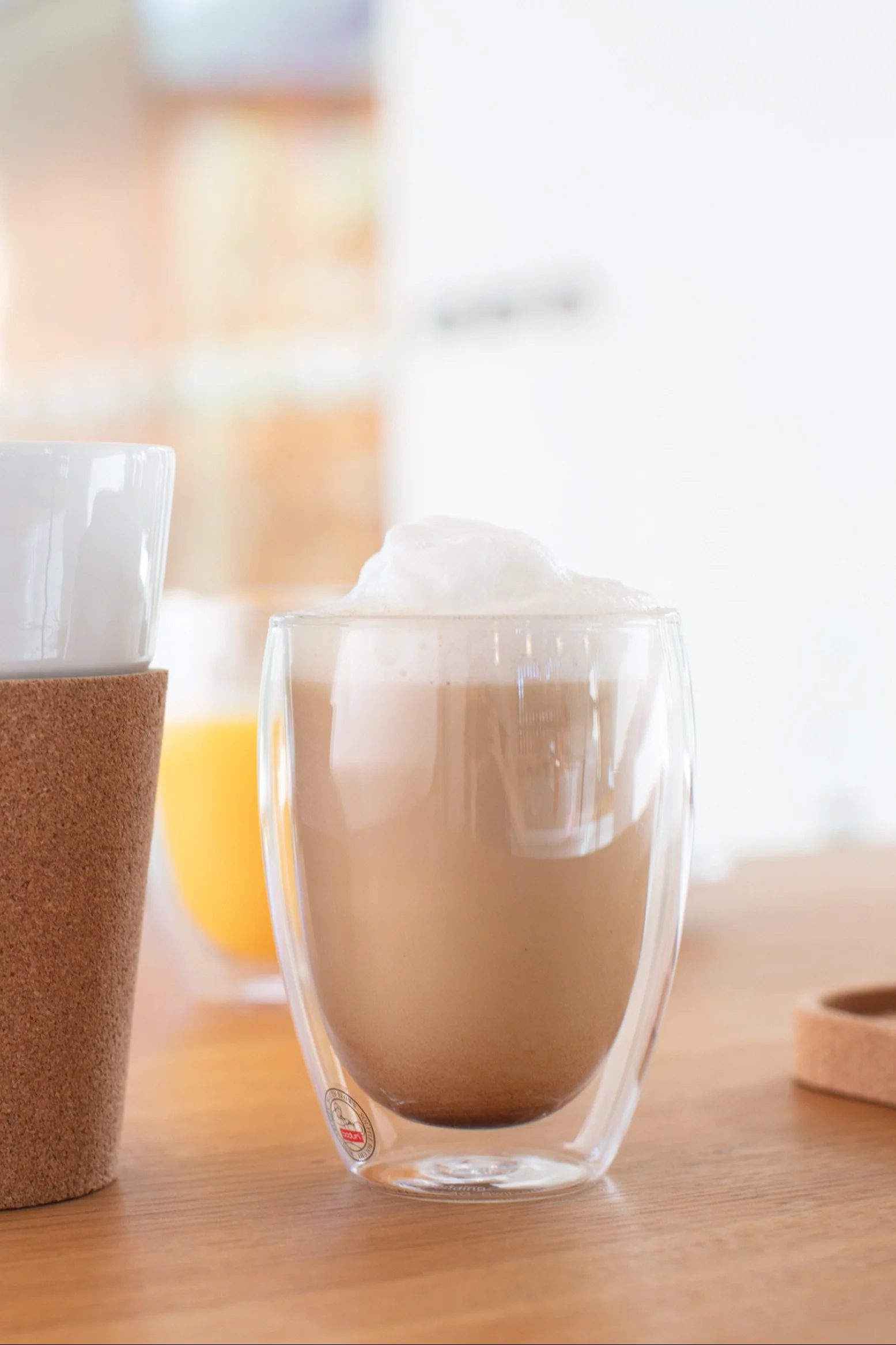 Foamy latte in a double-walled clear glass mug on a wooden table, with another mug with a cork sleeve to the left and an orange drink in the background.