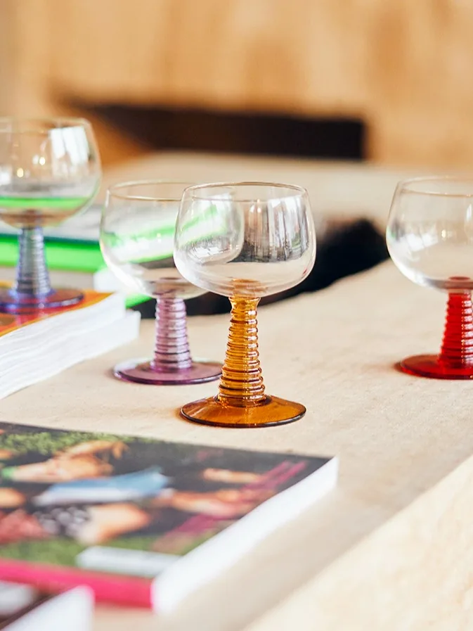 Several clear glass goblets with colorful ribbed stems (amber, purple, red) on a light surface next to stacks of magazines.