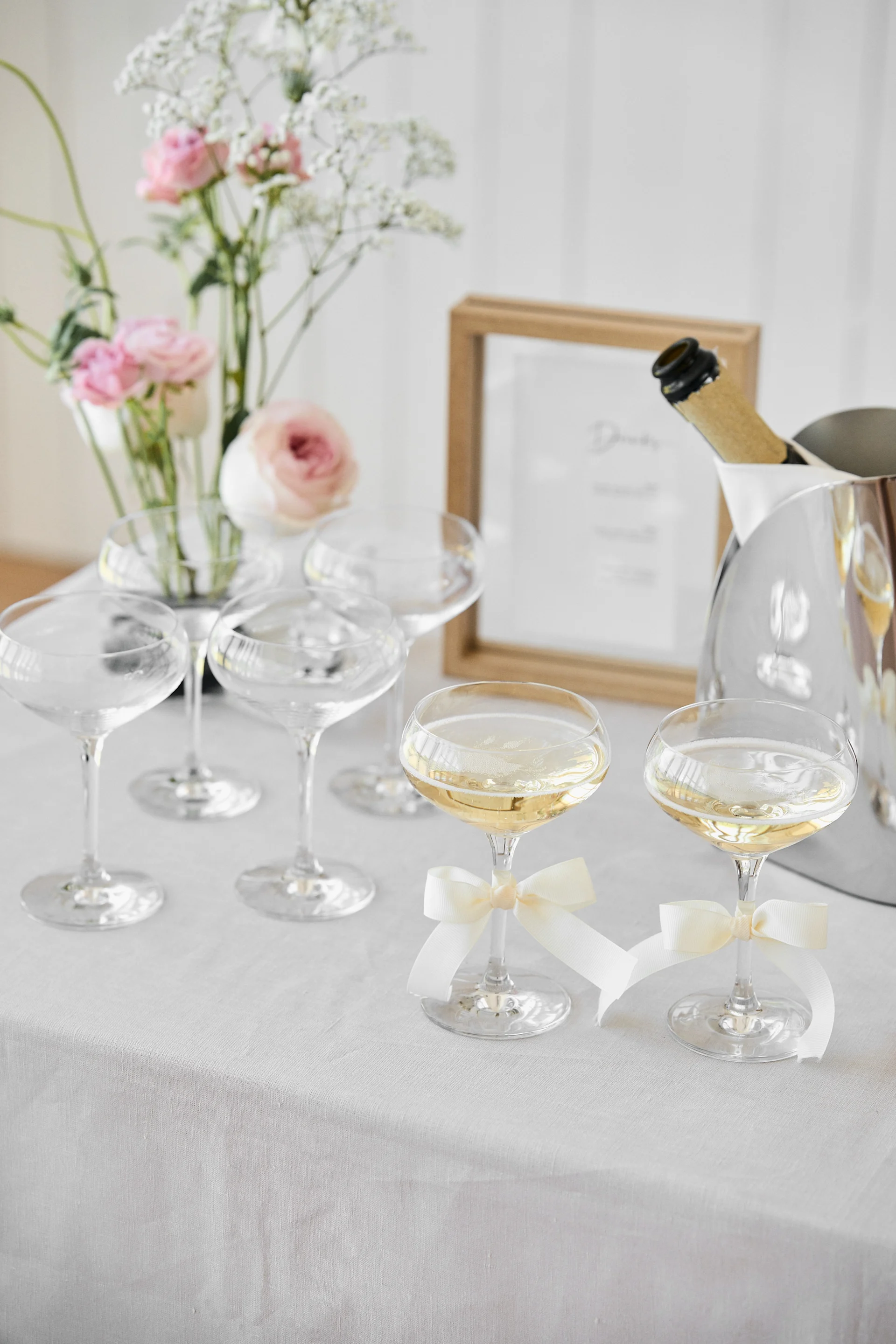 Celebration table with champagne coupe glasses, two with sparkling wine and white bows, a cooler, and a floral arrangement.