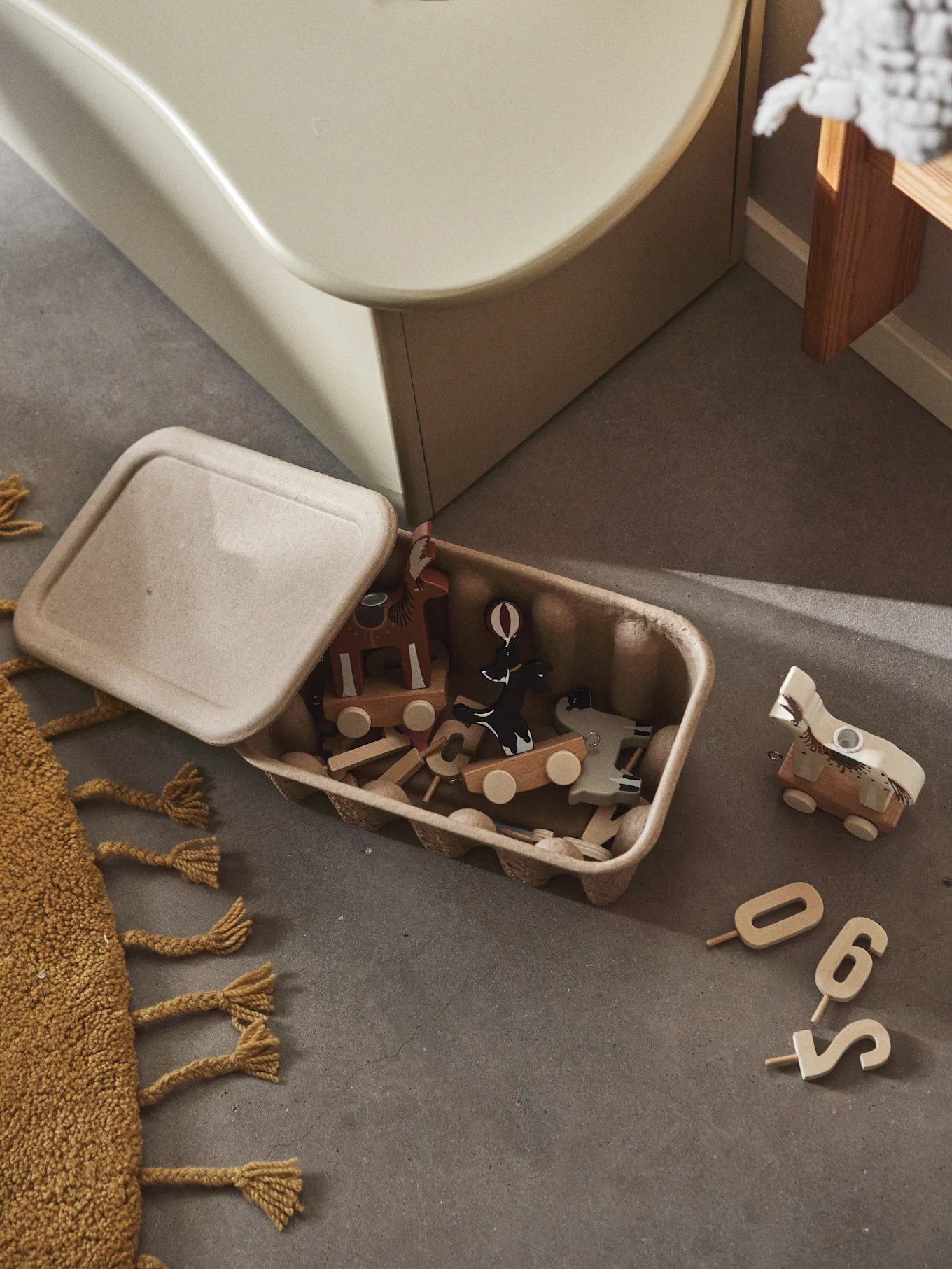 Wooden animal toys and blocks spill from an open, light brown container onto a concrete floor. Scattered wooden numbers and a yellow rug are visible.