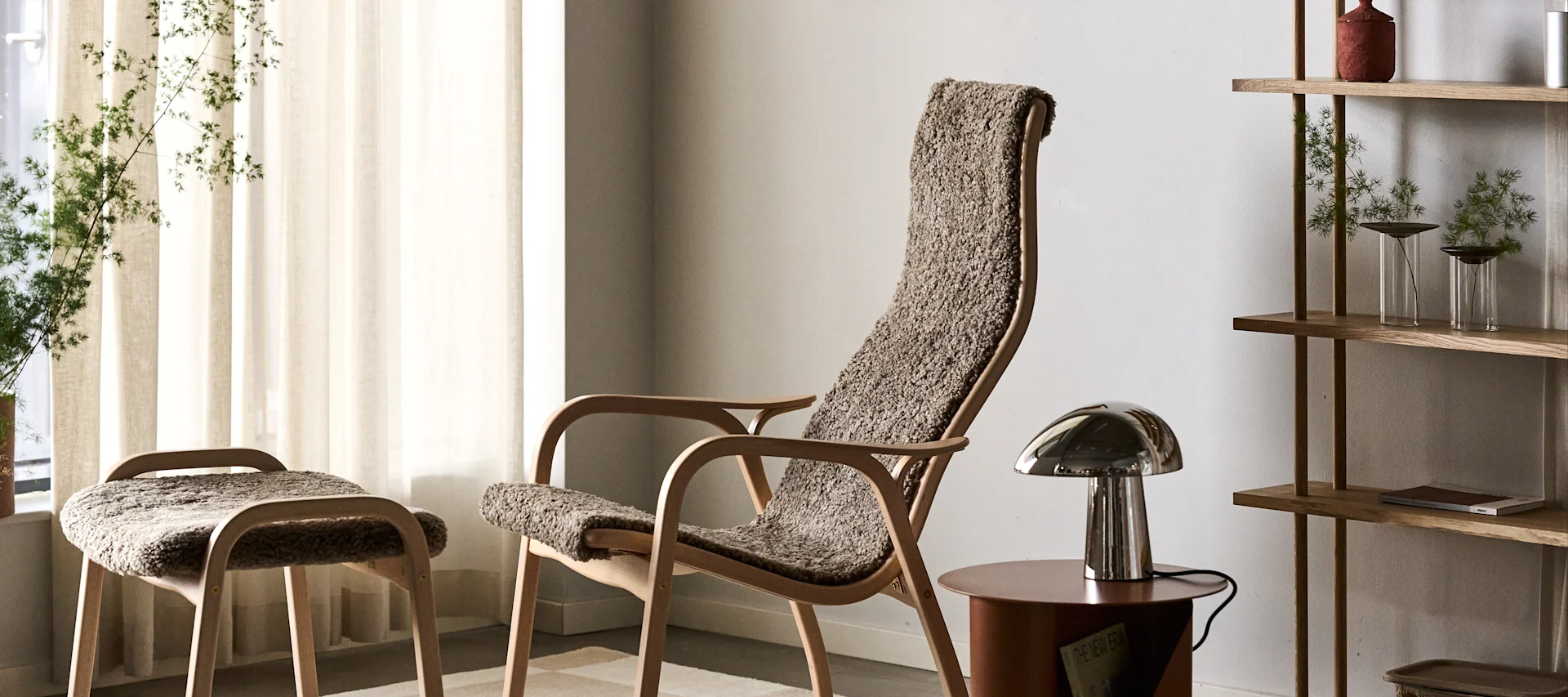A light wooden armchair and stool with textured brown upholstery, metallic table lamp, and wooden shelf with plants.