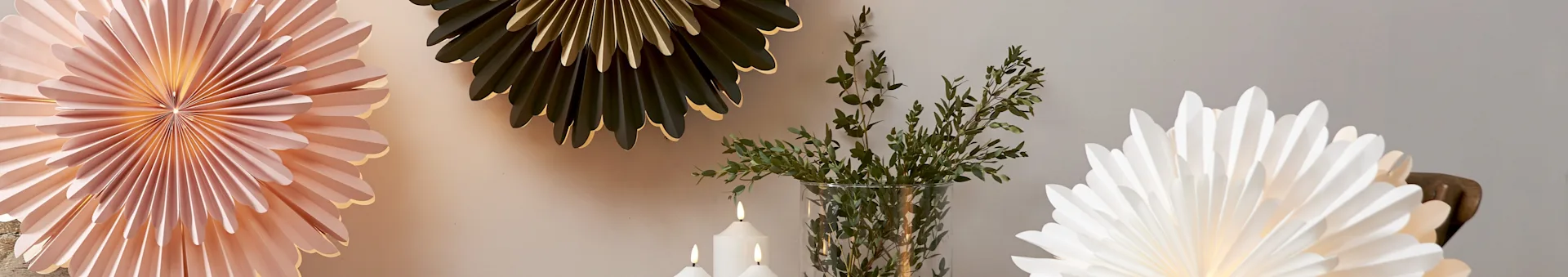 Three decorative folded paper fans in pink, dark green, and white, with lit candles and green branches in a clear vase.