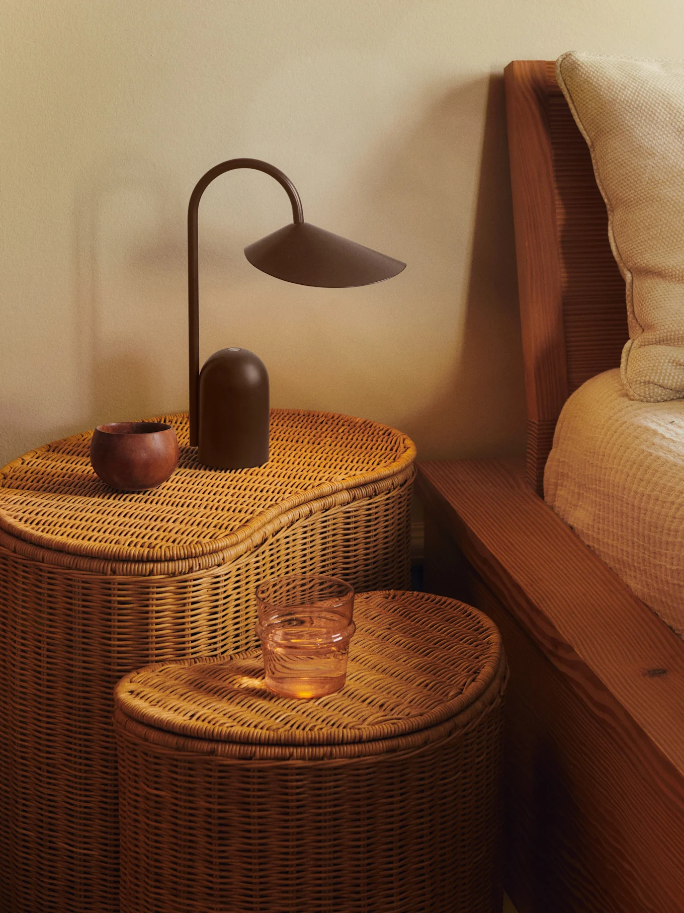 Two woven rattan bedside tables with a dark brown lamp, a wooden bowl, and a glass of water.