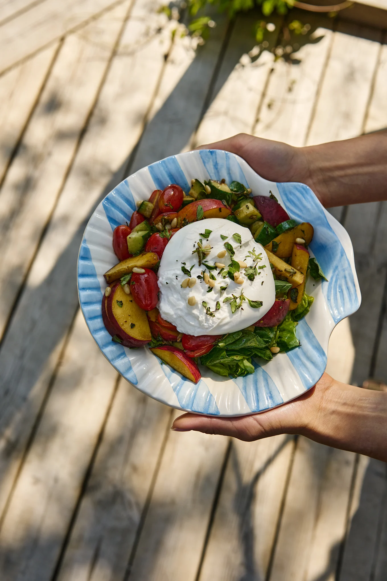 Hands hold a burrata salad with tomatoes, peaches, cucumber, and greens in a striped shell-shaped bowl.