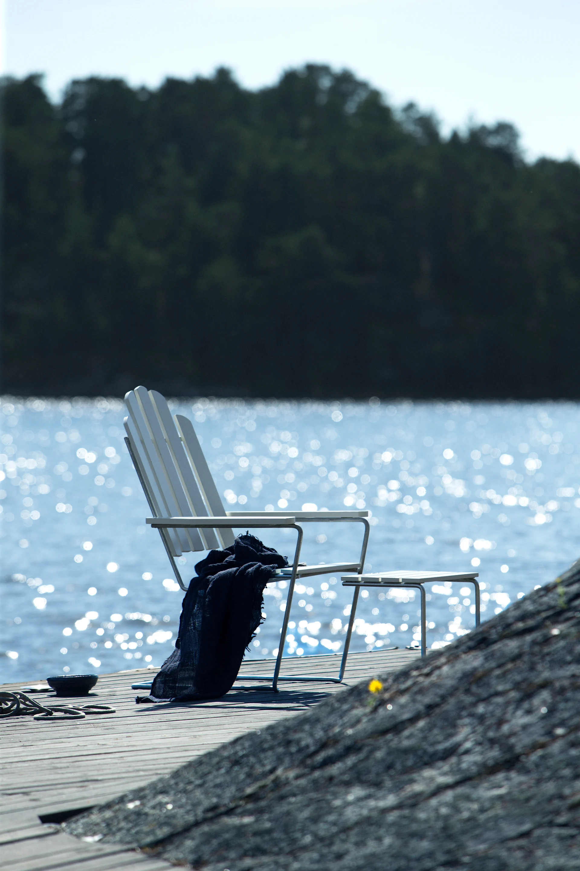 White slatted chair with dark blue blanket on a wooden dock by sparkling water, with trees in the background.