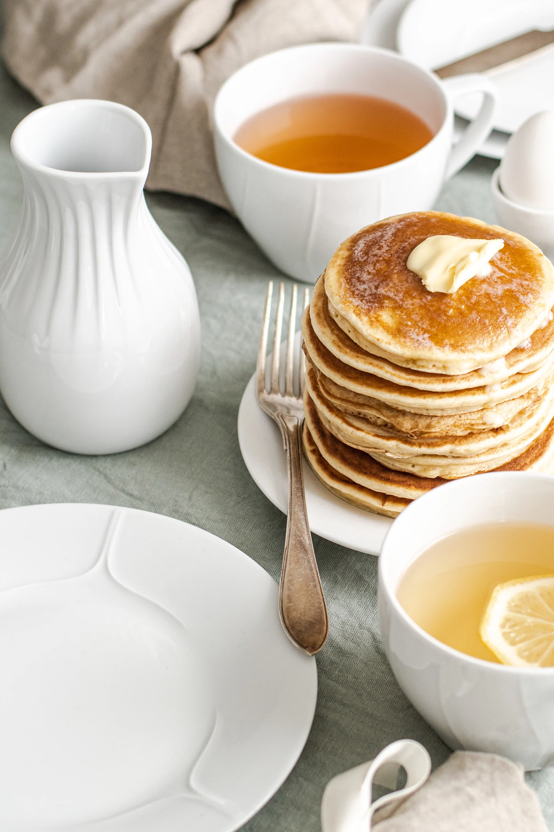 Stack of pancakes with melting butter, served with tea and a pitcher on a light green tablecloth.