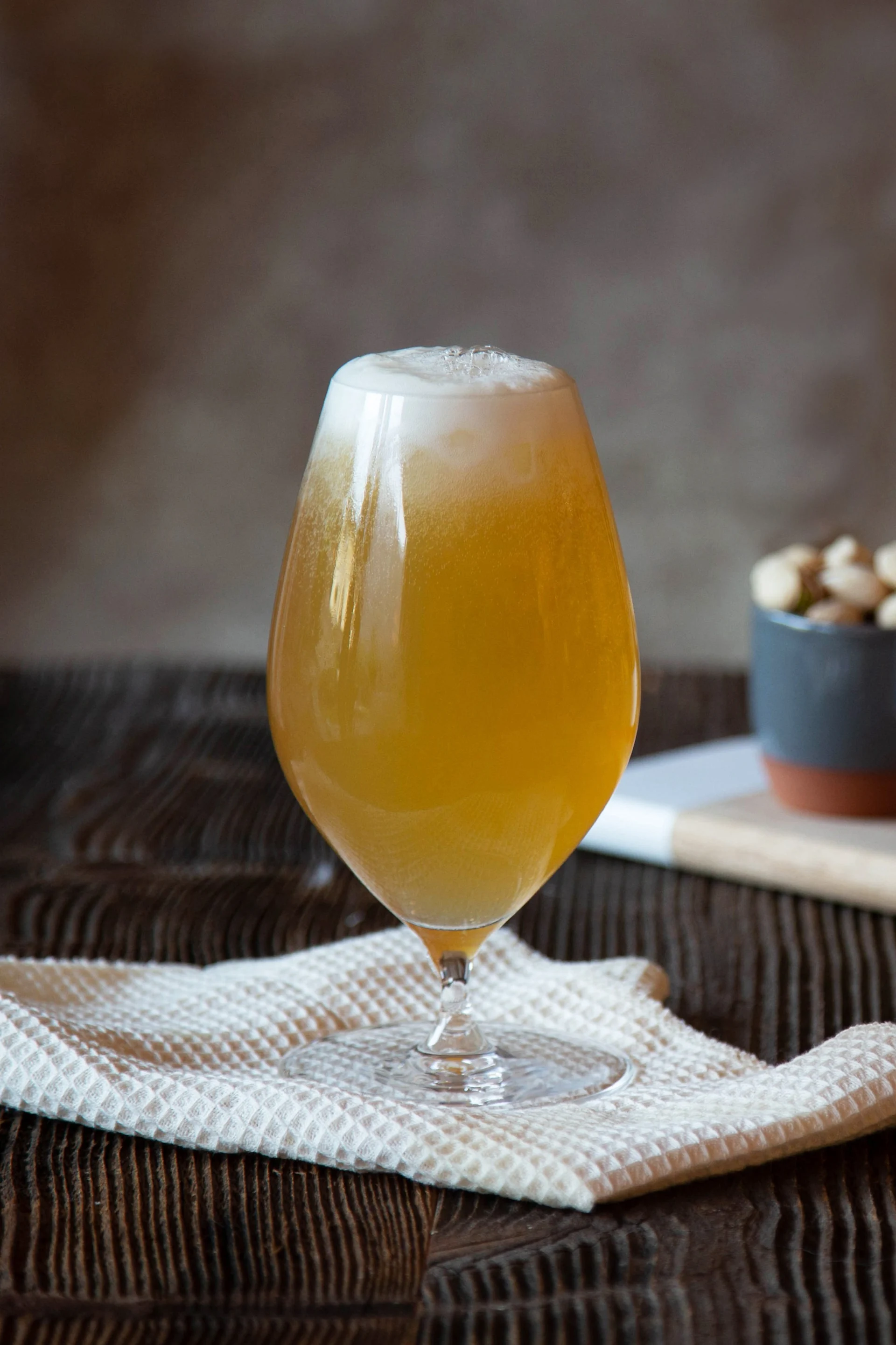 A glass of frothy amber beer on a white cloth, with bowls of nuts and breadsticks in the background on a wooden board.