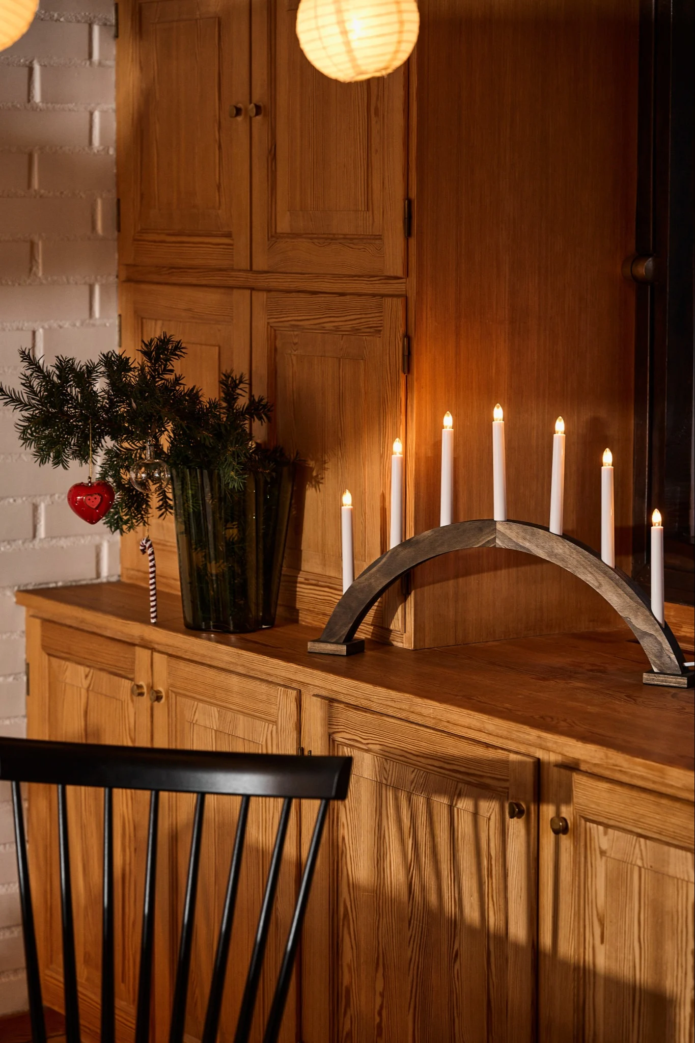An arched Advent candle holder in dark stained wood stands on a wooden chest of drawers, next to a vase filled with spruce branches.