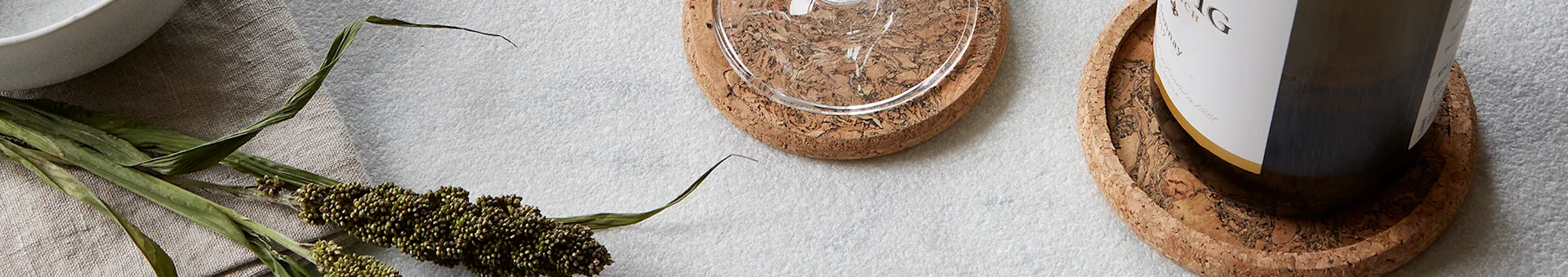 Two cork coasters, one holding a glass and the other a wine bottle, next to dried green stalks on a linen cloth.