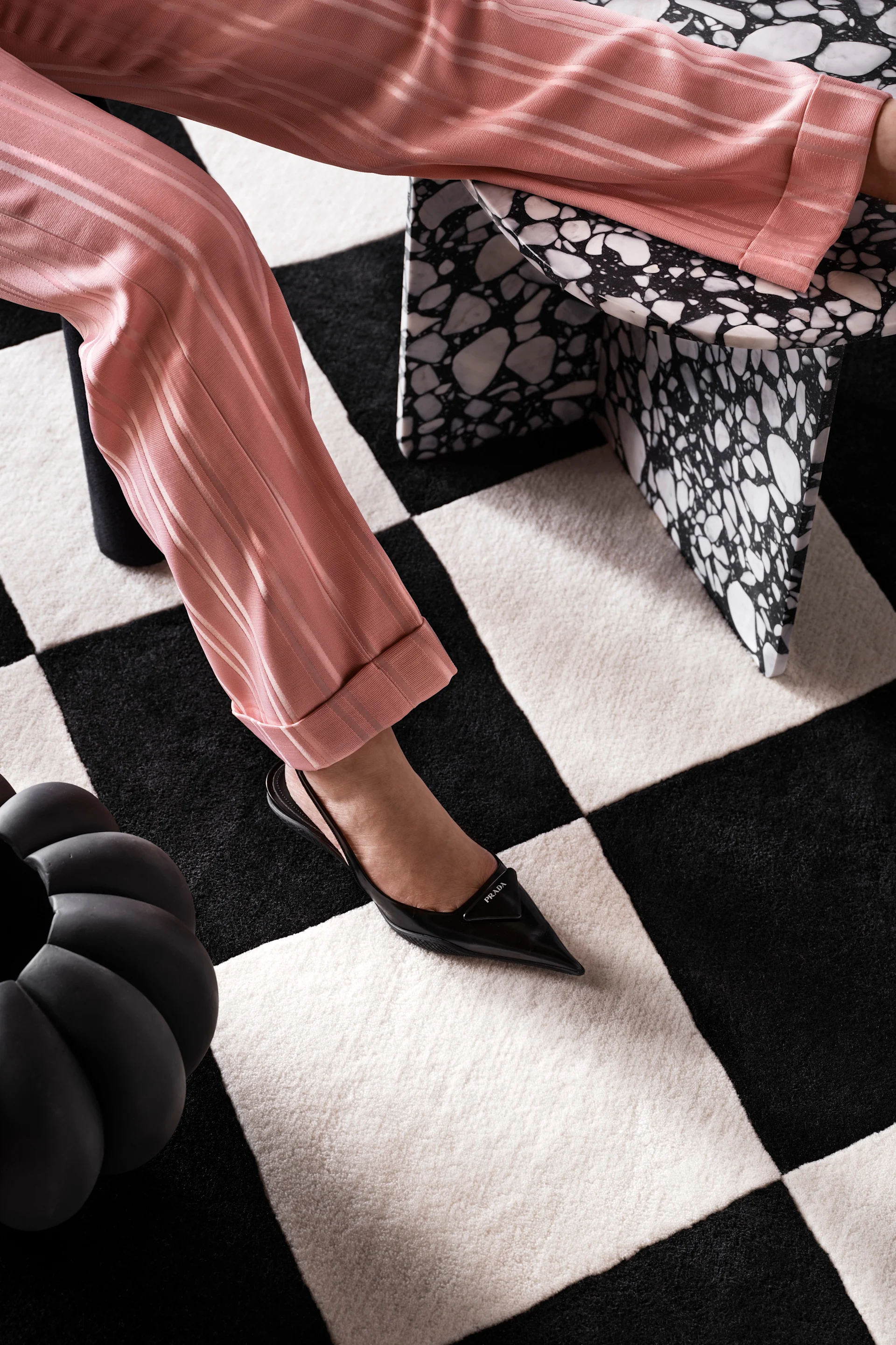 Person in pink striped pants and black pointed-toe slingback heels with triangular detail, sitting on a terrazzo stool on a checkerboard rug.