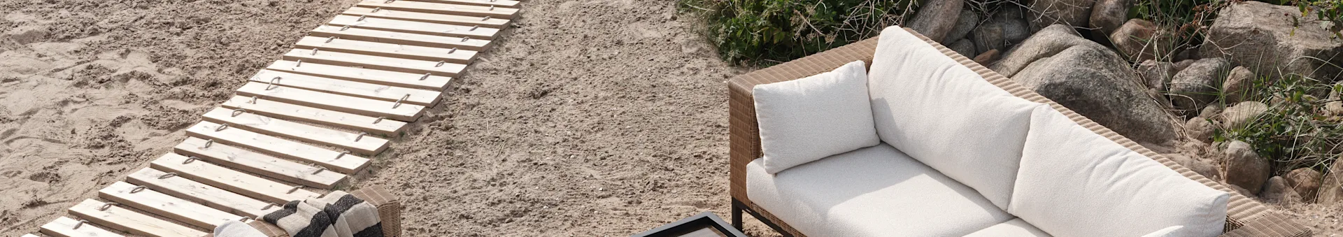A wooden walkway on sand leading to a rattan outdoor sofa with white cushions and a striped blanket, set against rocks and green plants.