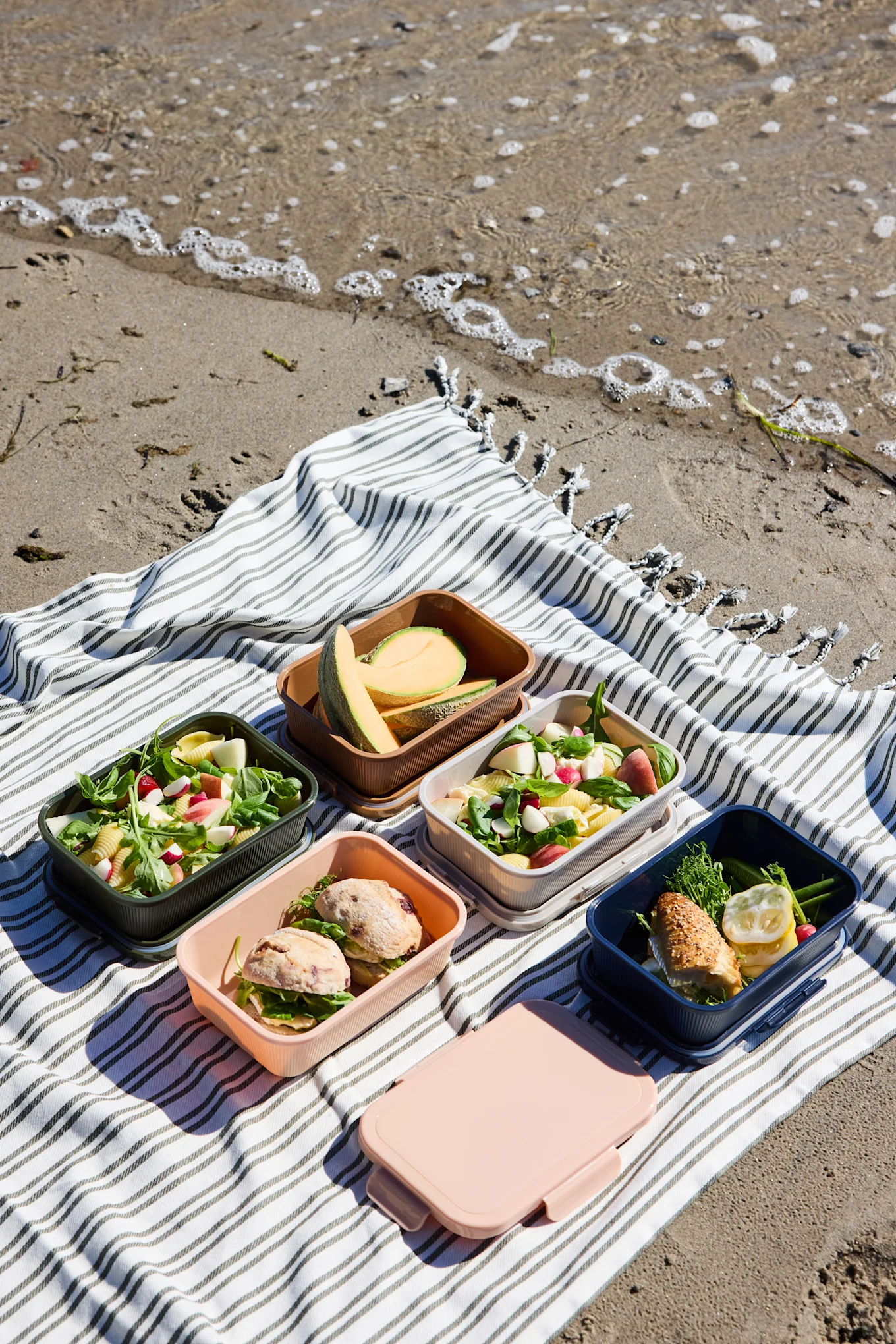 Five open colorful lunchboxes with salads, sandwiches, and melon, plus a closed pink box, on a striped beach blanket.