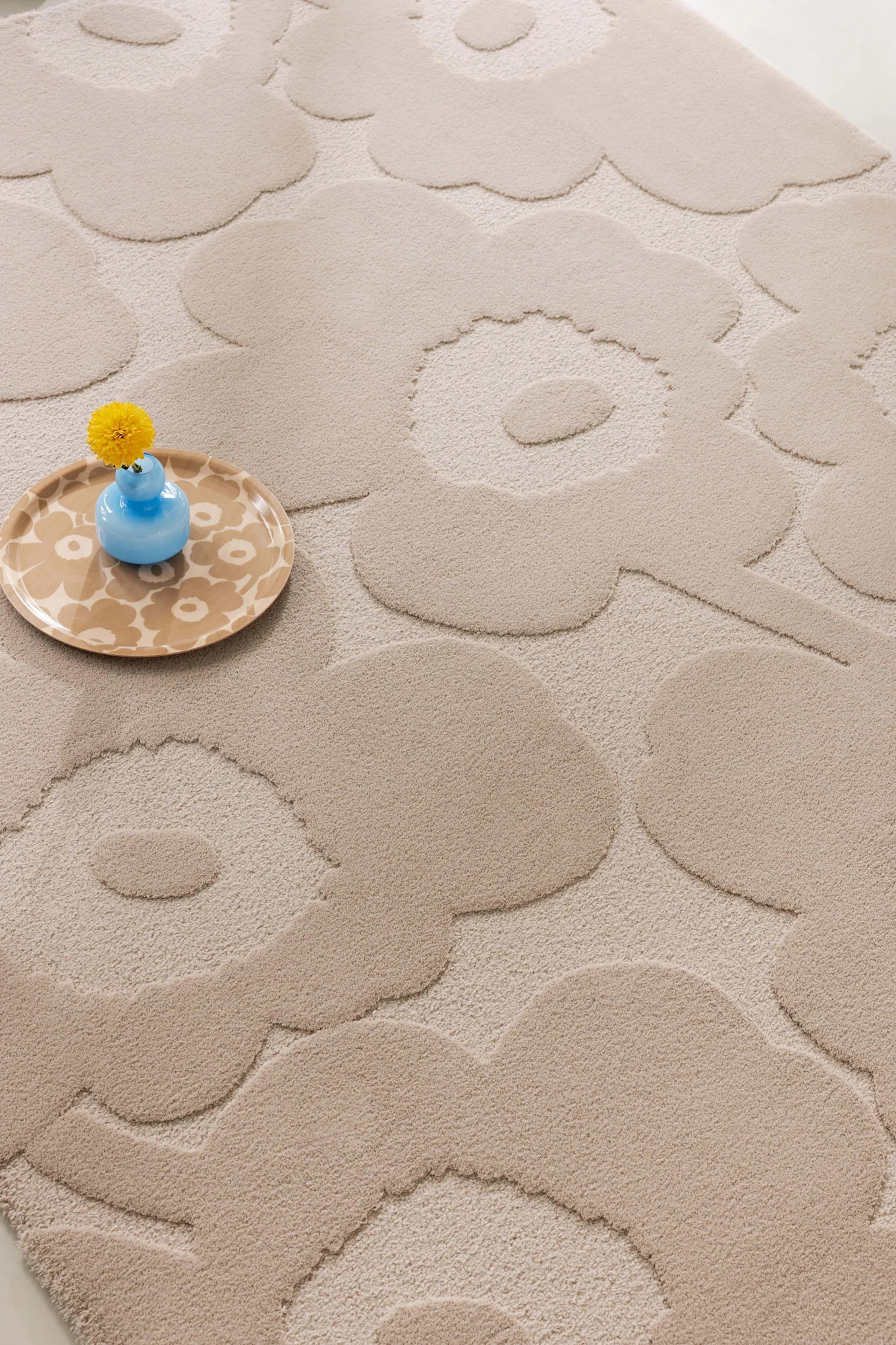 Beige rug with a raised floral pattern, featuring a light blue vase with a yellow flower on a patterned tray.