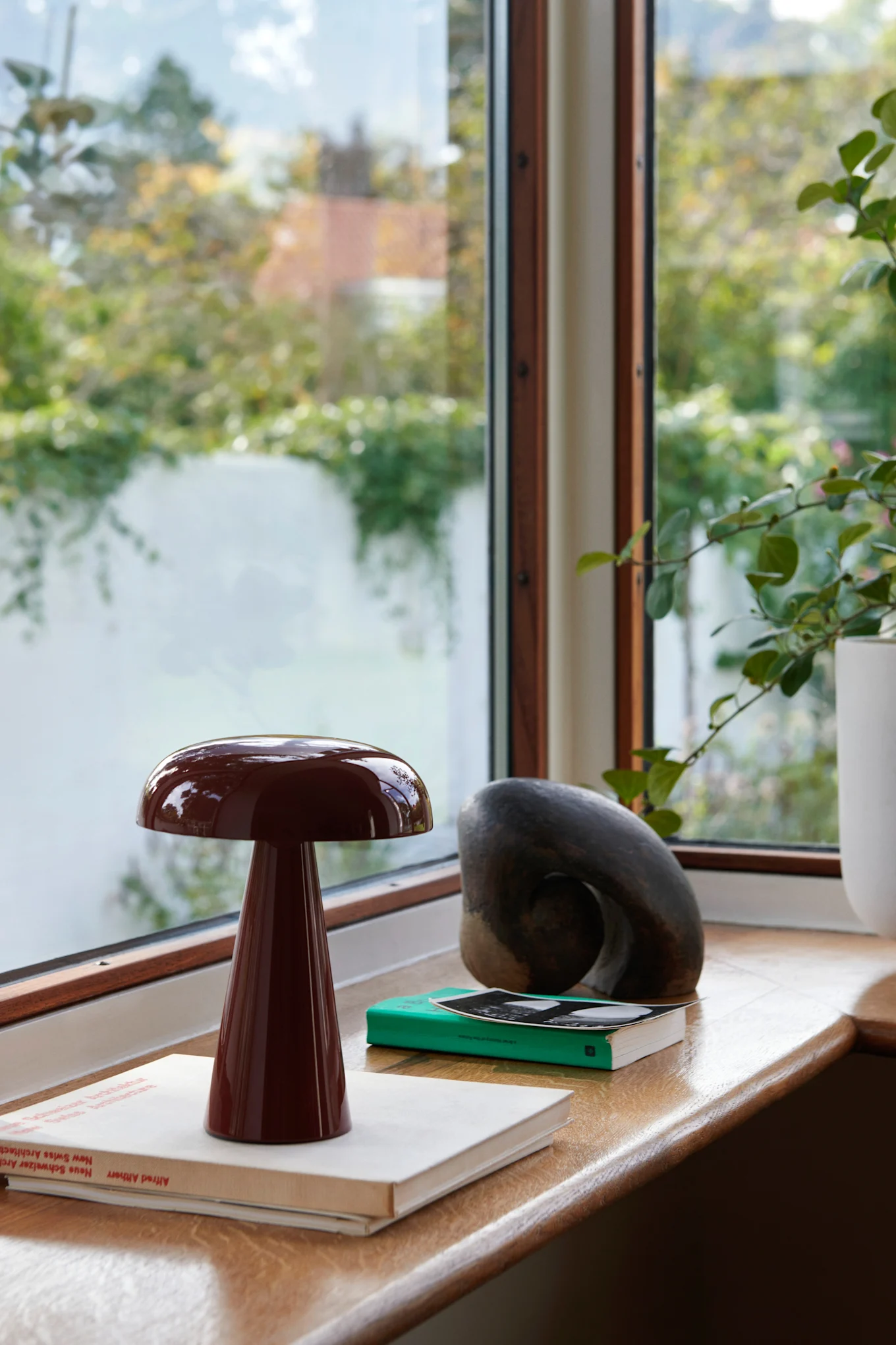 Dark red mushroom lamp on a windowsill with books and a sculpture, green foliage visible outside.