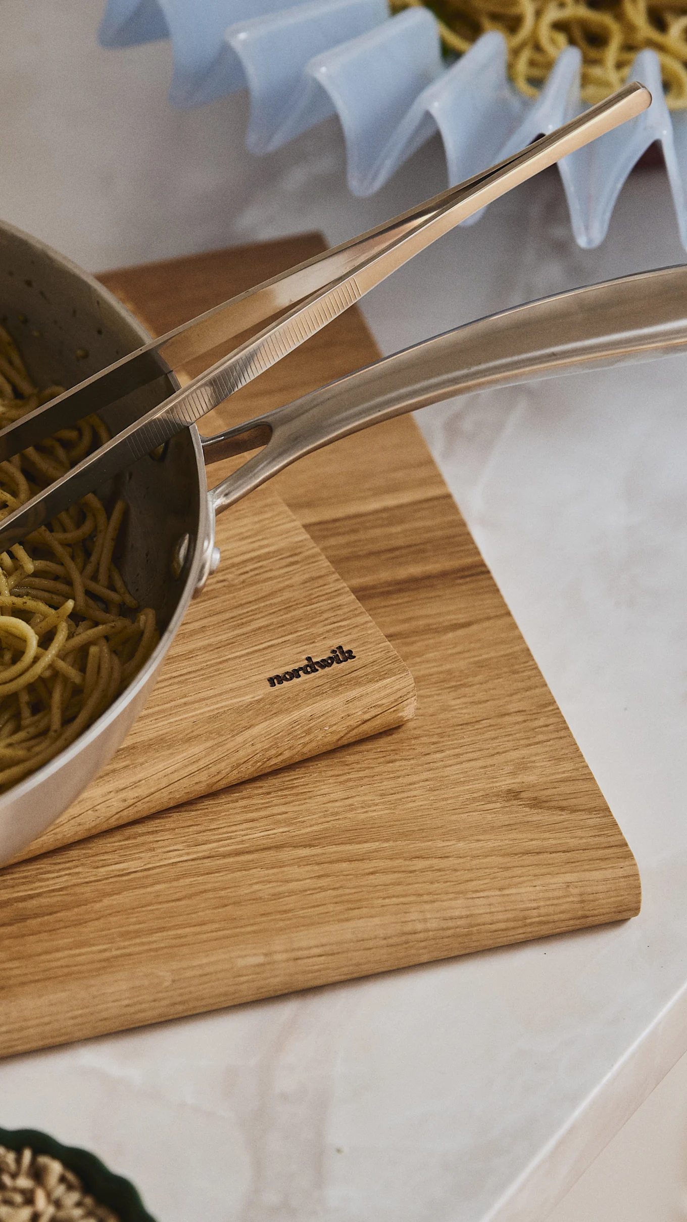 Close-up of pesto pasta in a metal pot with tongs, resting on two wooden cutting boards. One board has 'nordwik' engraved.