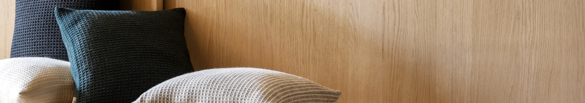 Three waffle-knit pillows in dark blue-grey and light beige against a light wooden wall.