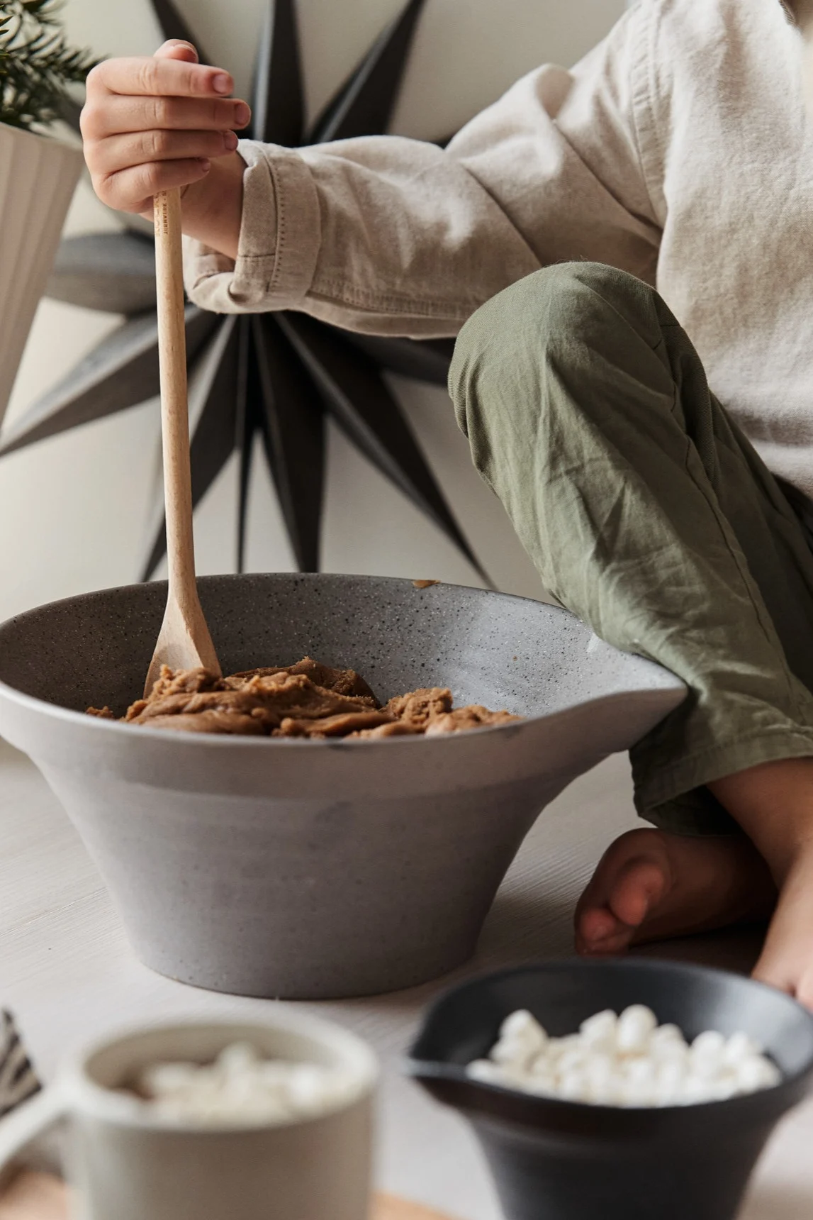 Child mixing brown batter in a grey bowl with a wooden spoon, small white treats in foreground.