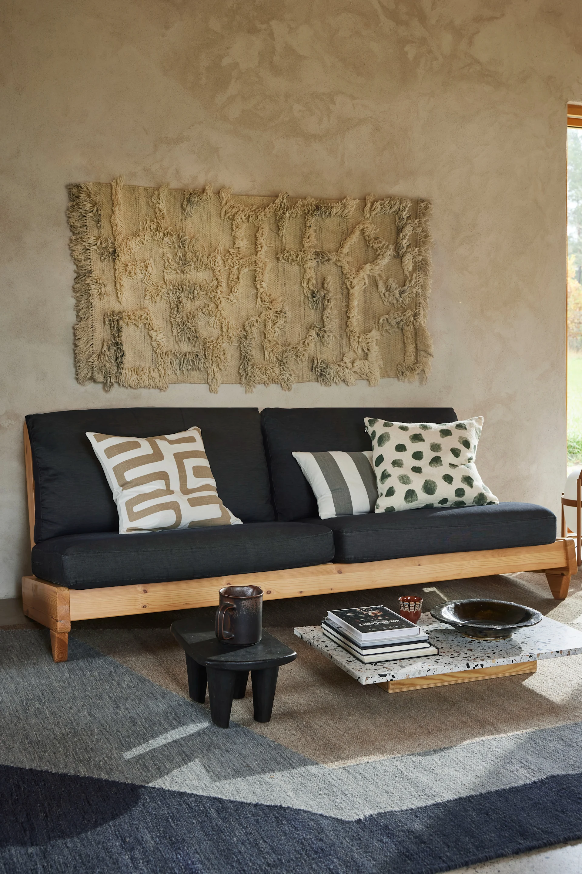 Cozy living room with dark grey sofa, patterned pillows, textured wall art, and multi-toned rug. Coffee table.
