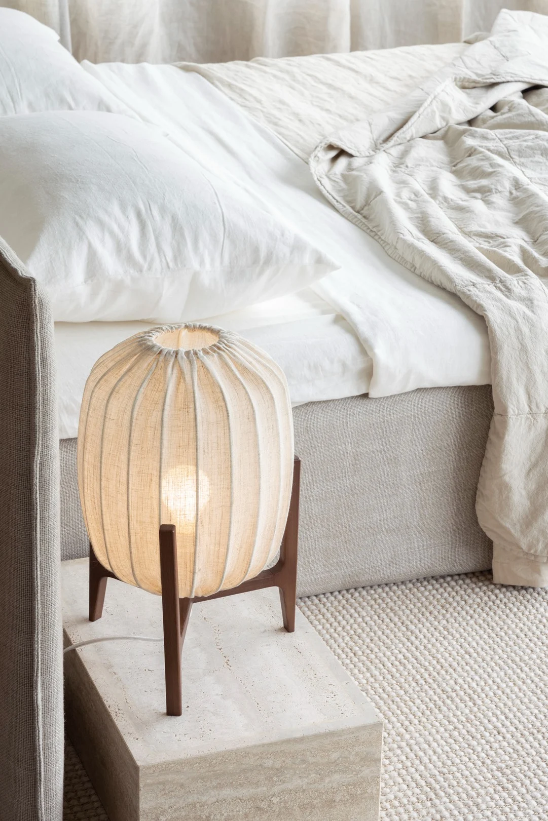 A bedside table with a glowing pleated fabric lamp, beside a bed with white sheets and a beige comforter.