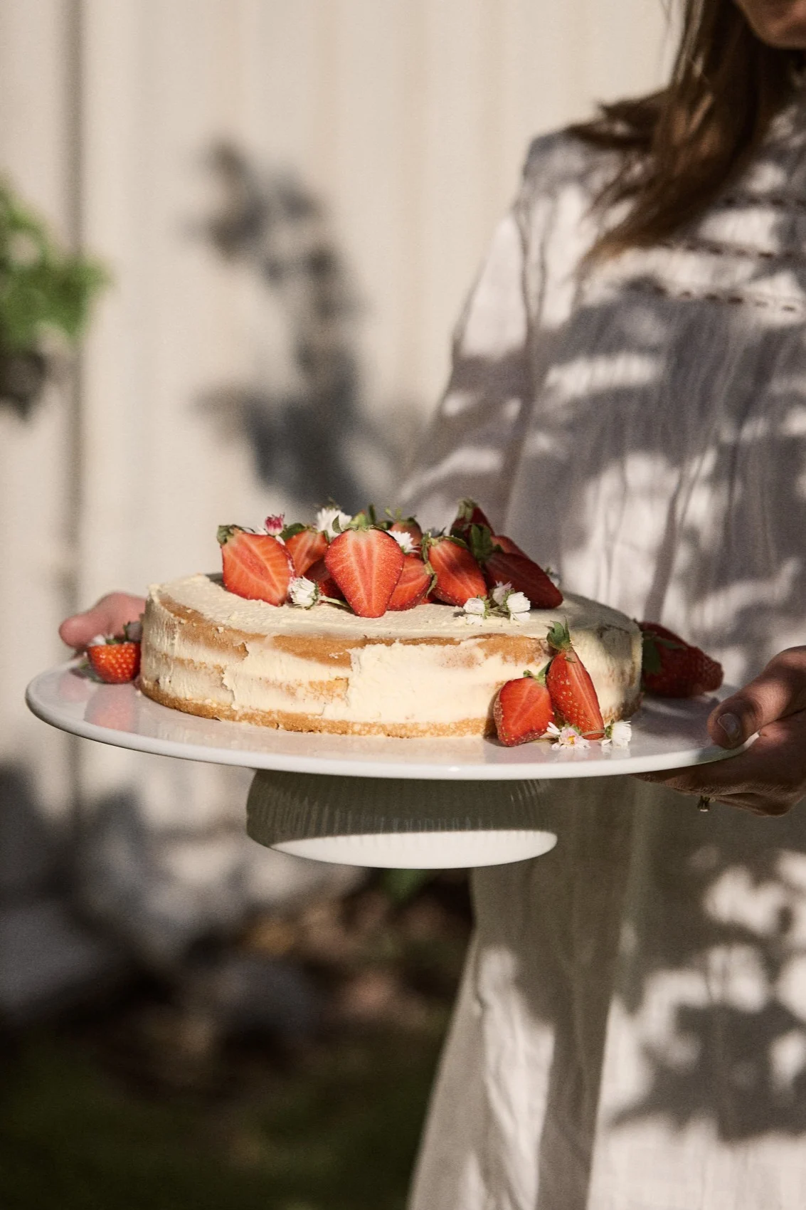 Person holding a round cake topped with fresh halved strawberries and small white flowers on a white cake stand outdoors.