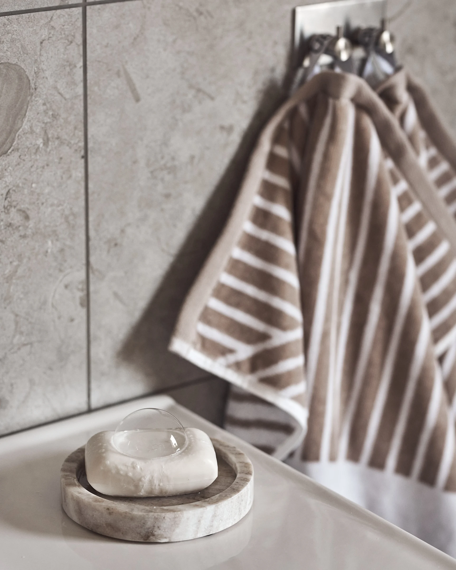White soap in a marble dish on a white sink, with brown and white striped towels hanging on a tiled wall.