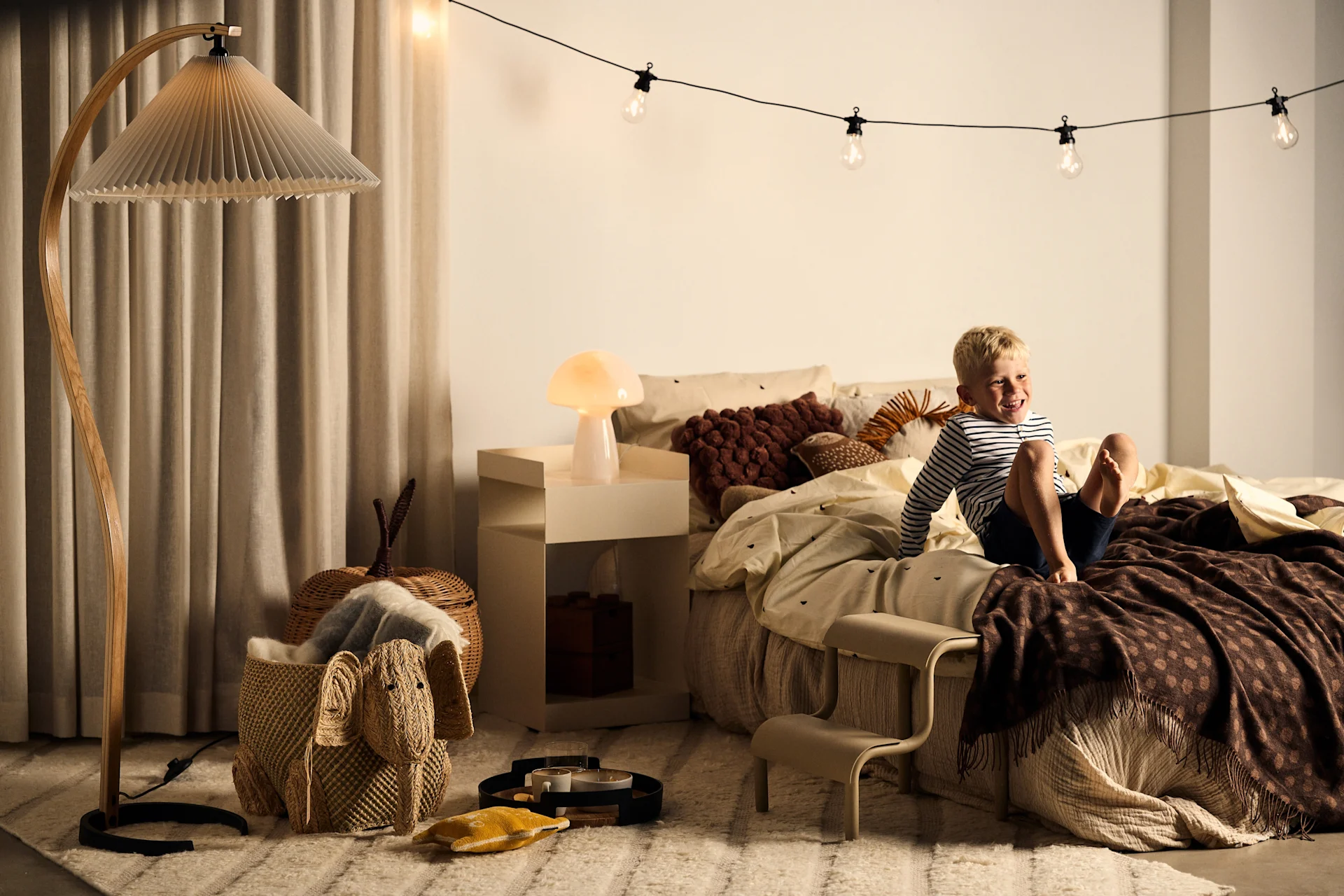 A smiling boy sits on a bed with a brown polka-dot blanket, next to a pleated floor lamp and a mushroom table lamp.
